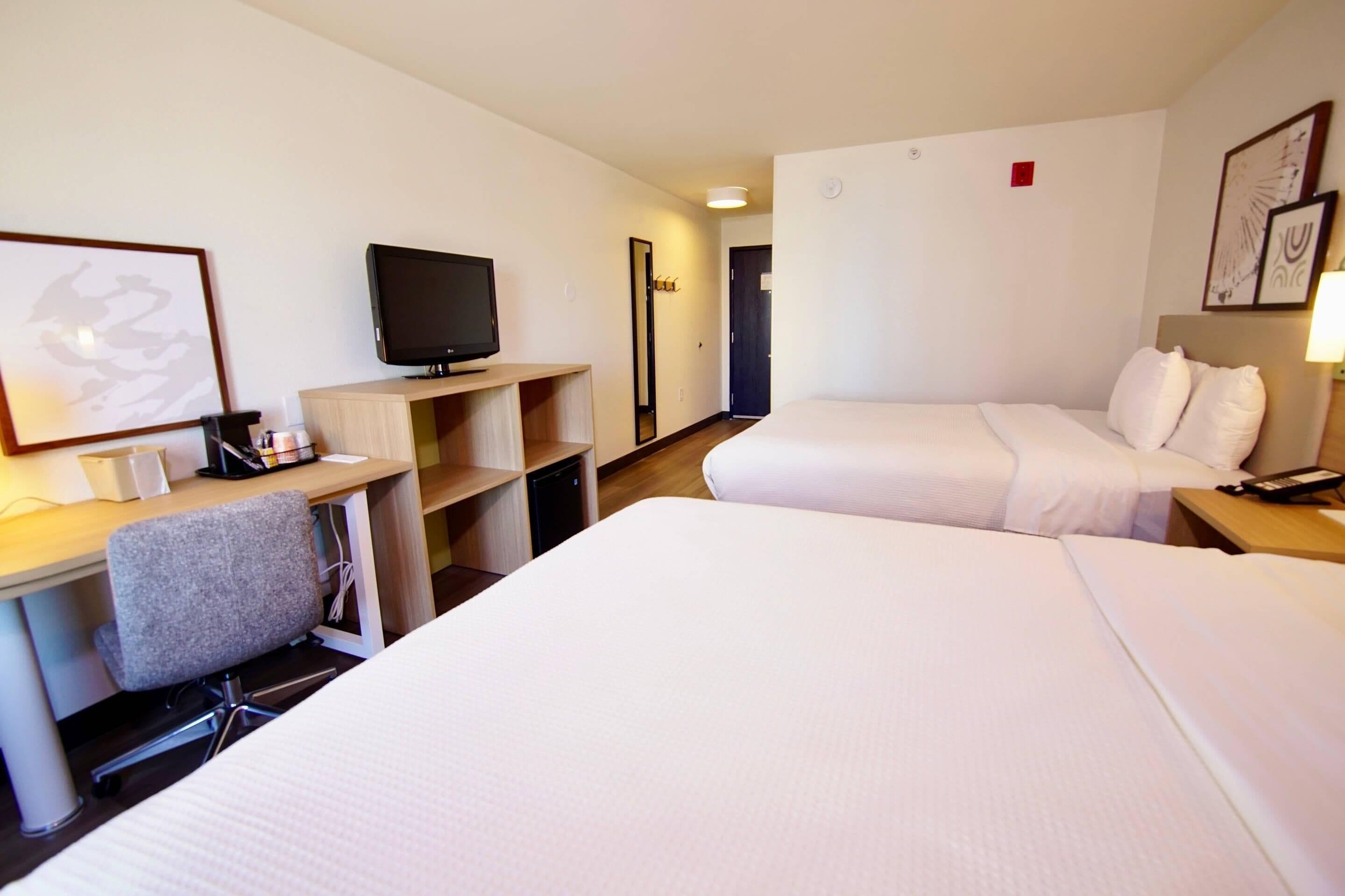 Una habitación de hotel con dos camas, un escritorio con silla, un televisor y un espejo.