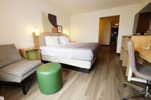 Habitación de hotel con cama, escritorio, silla, reposapiés verde y televisor montado en la pared.