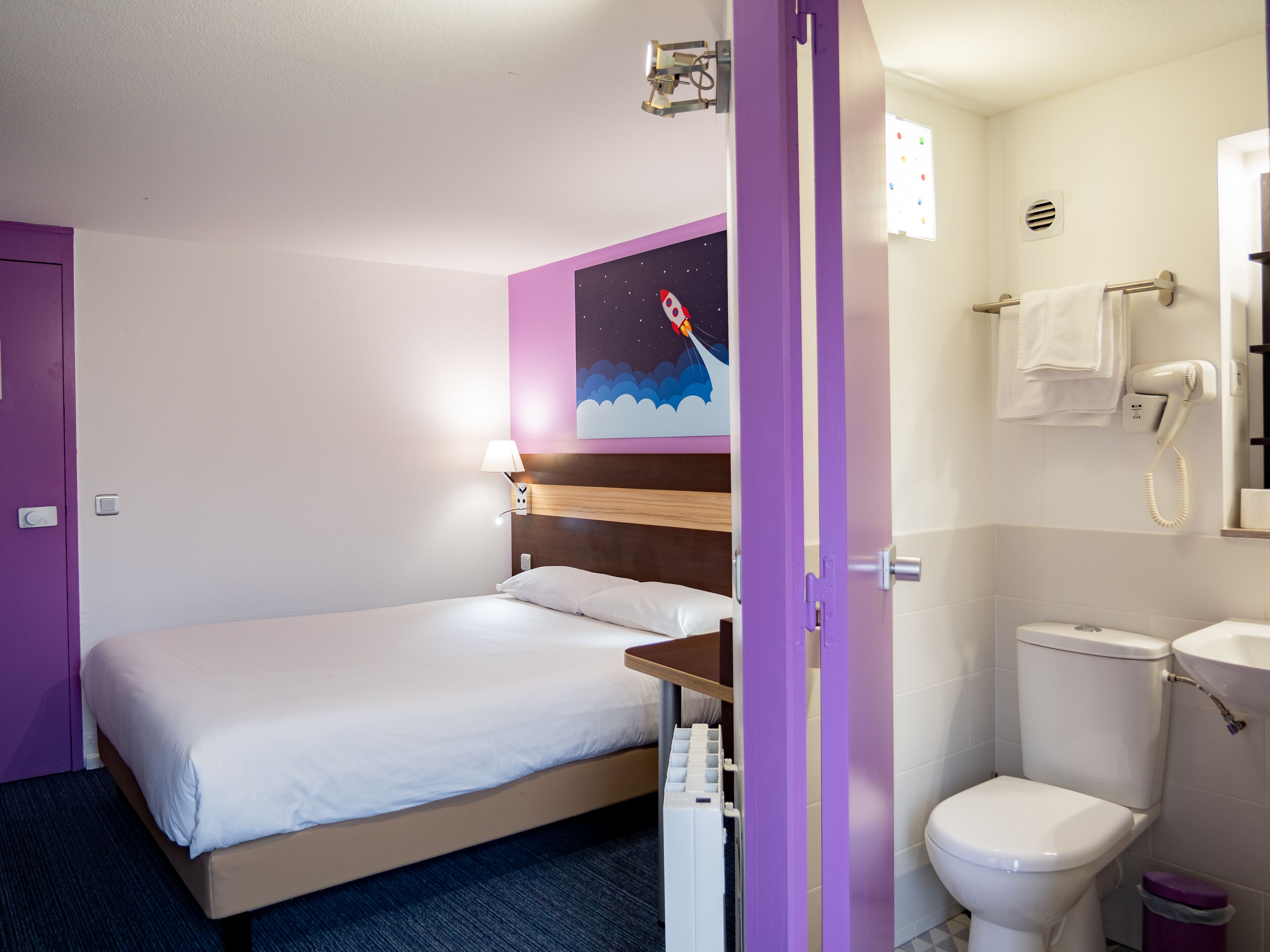 Una habitación de hotel con una cama, una mesita de noche, una lámpara de pared, un baño con inodoro, lavamanos y secador de pelo.