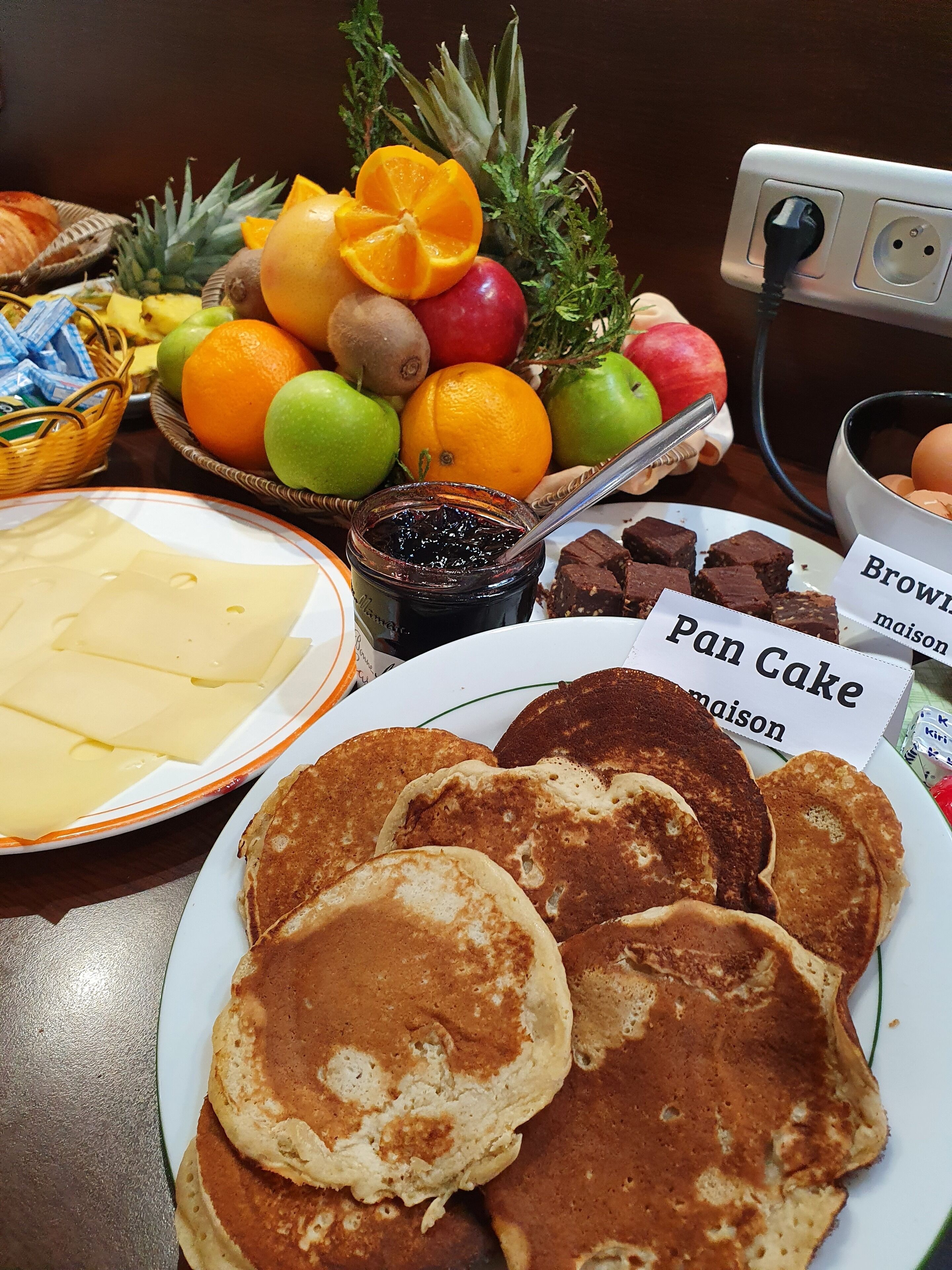 Une table de petit-déjeuner avec des crêpes, une assiette de fromage, un bol de fruits et des étiquettes indiquant « crêpes » et « brownies ».