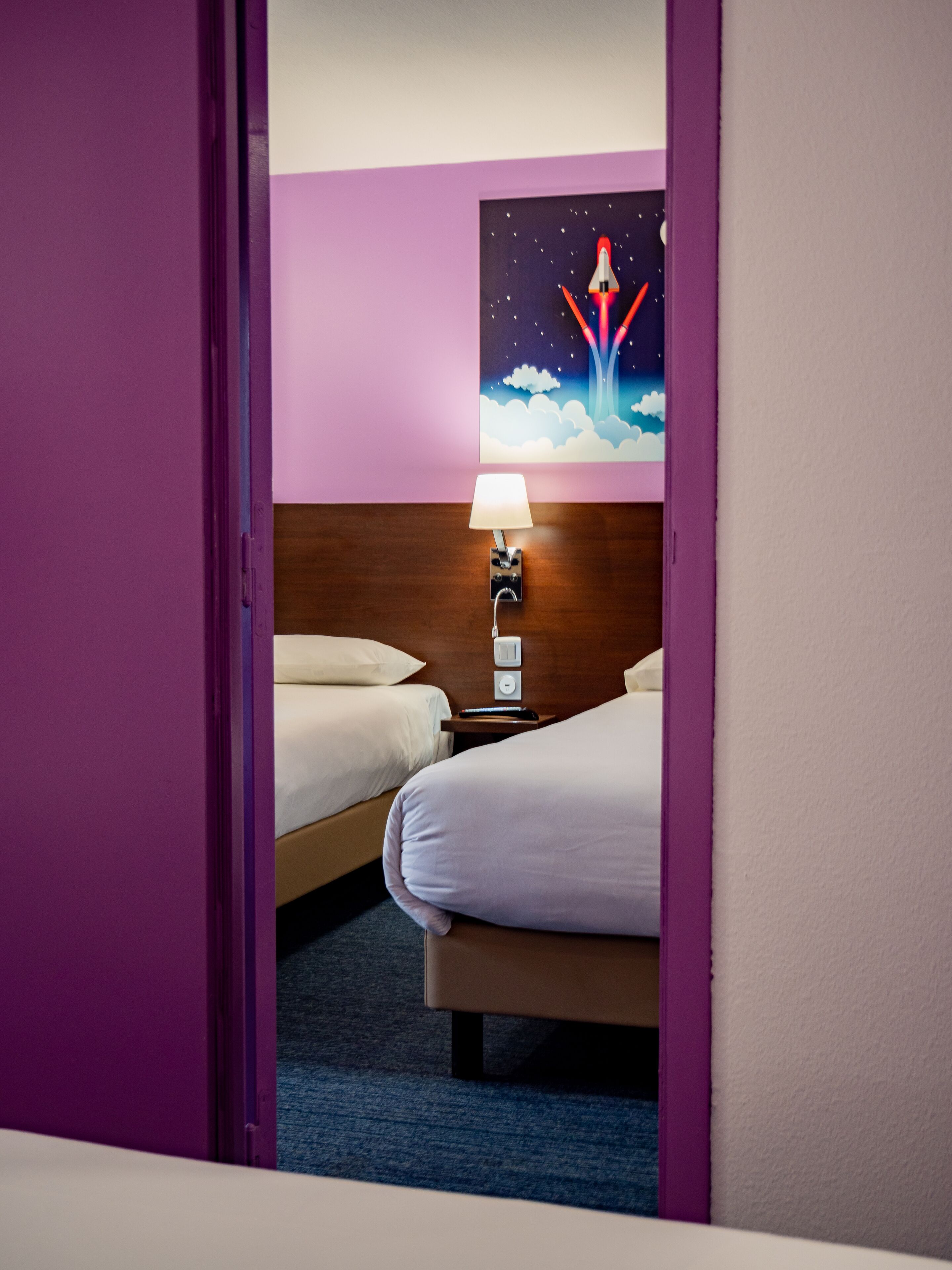 Habitación de hotel con dos camas, una mesita de noche con lámpara y un cuadro de un lanzamiento de cohete en la pared.