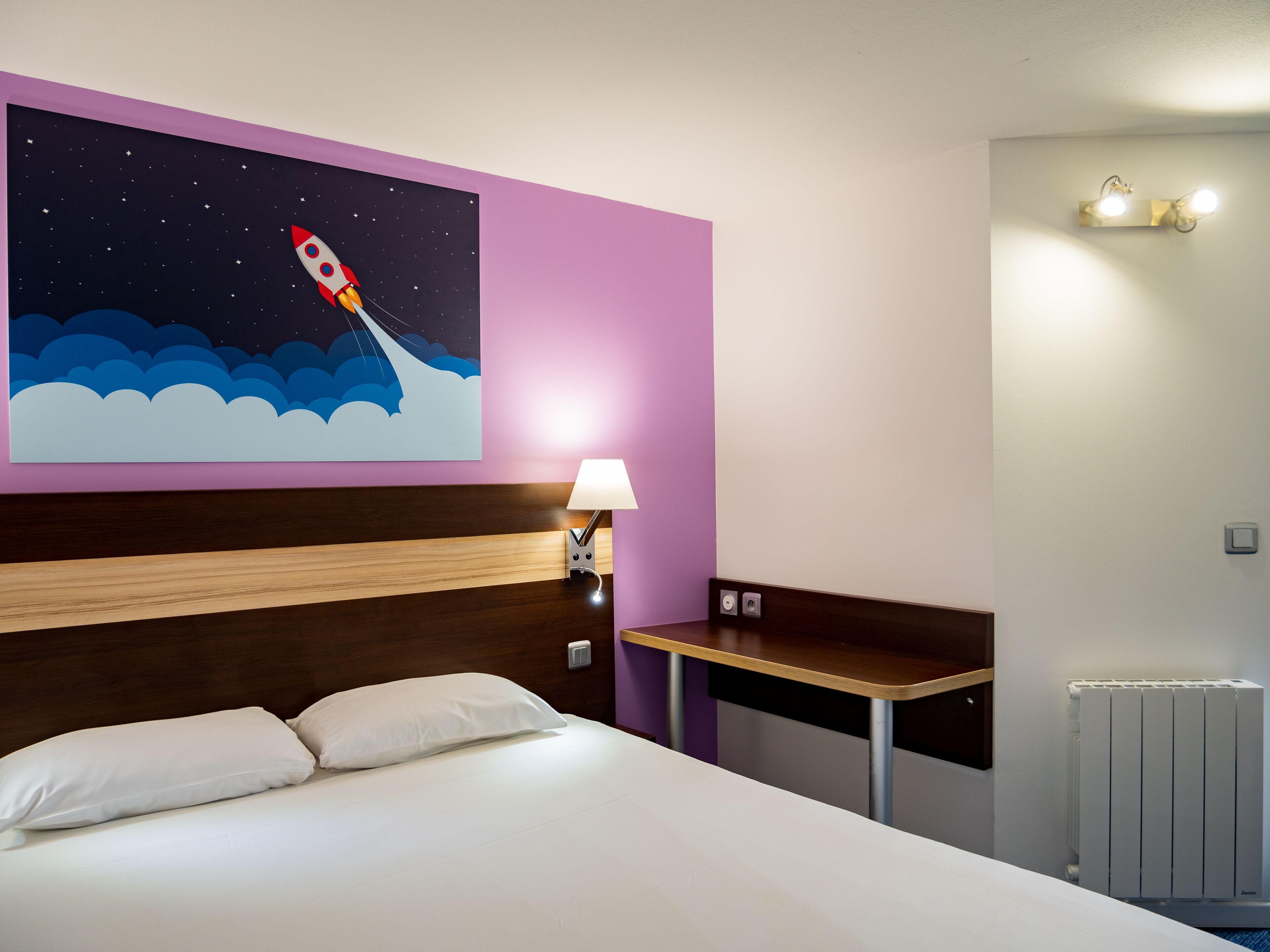 A hotel room with a bed, a night sky mural, a nightstand with a lamp, a bench, and a radiator.