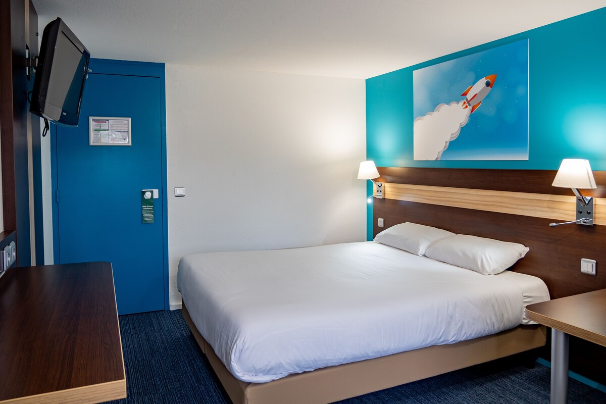A hotel room with a bed, a desk, a TV mounted on the wall, and a painting of a rocket.
