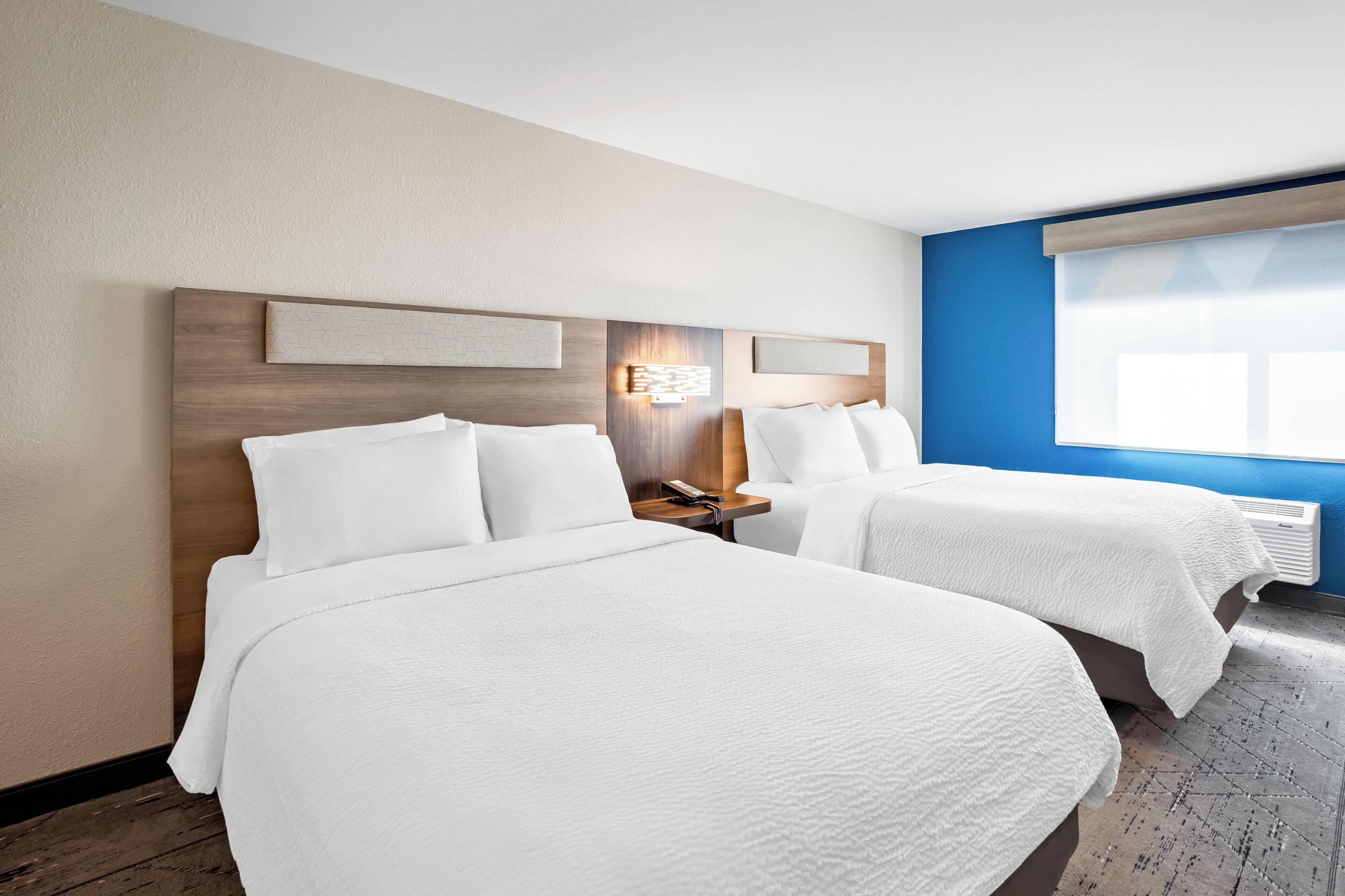 A hotel room with two beds, a nightstand with a lamp, and a window with blinds.