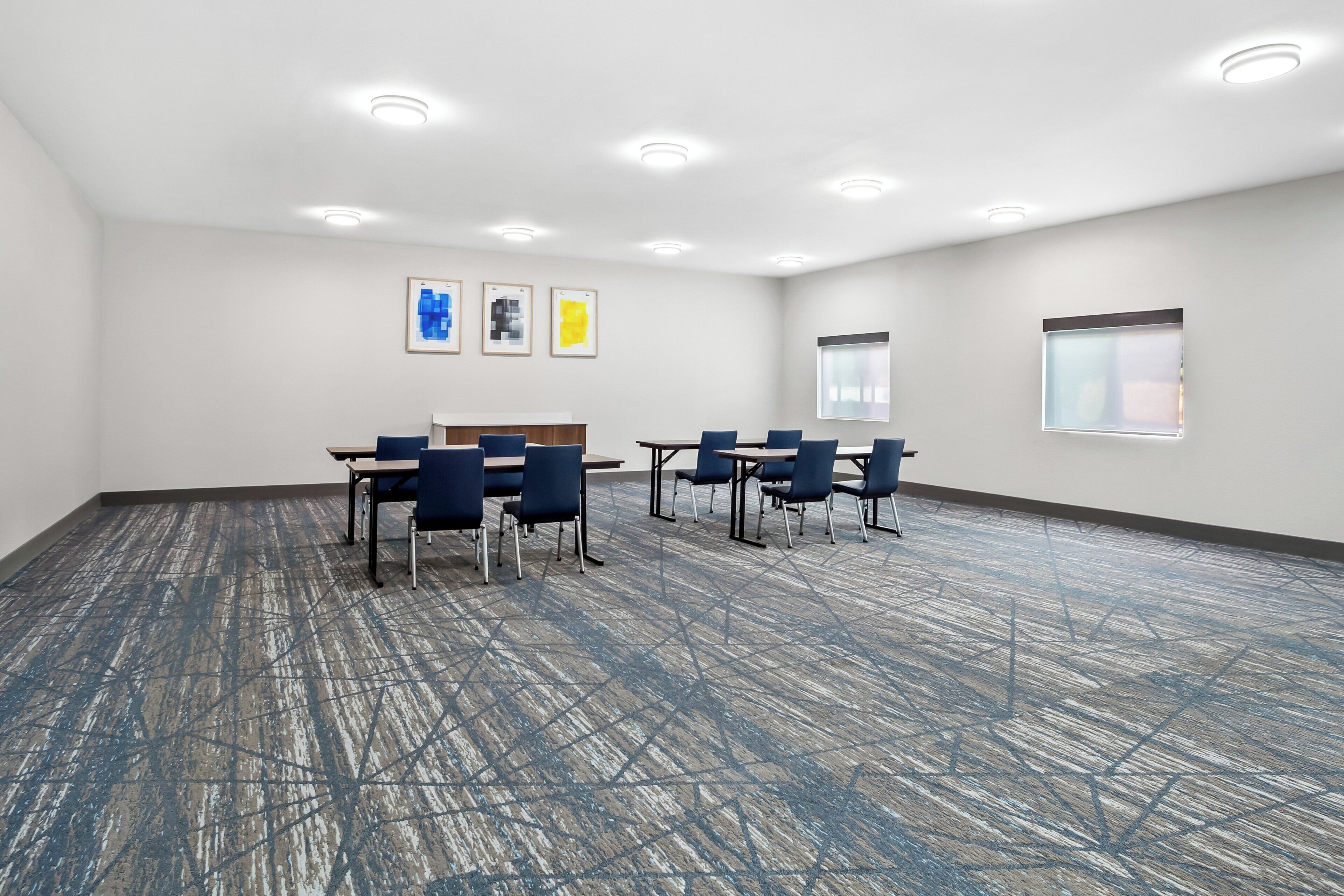 A conference room with a blue carpet, white walls, and a ceiling with recessed lighting. There are four tables with chairs, a projector screen, and framed artwork on the wall.