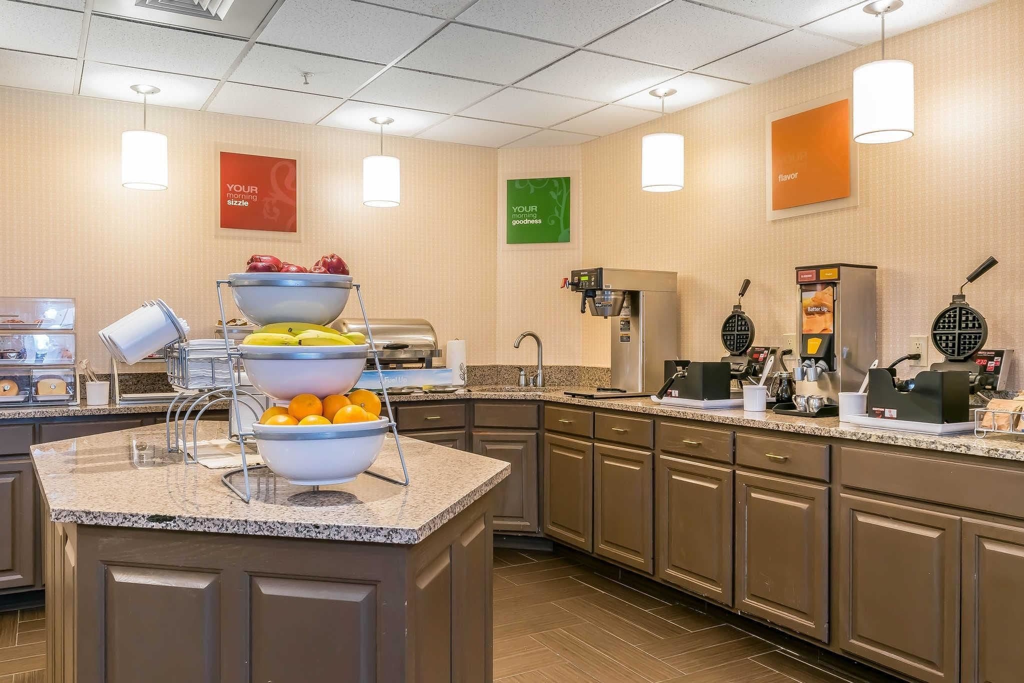 A hotel kitchen with a buffet setup, including a fruit bowl, waffle maker, and coffee station.