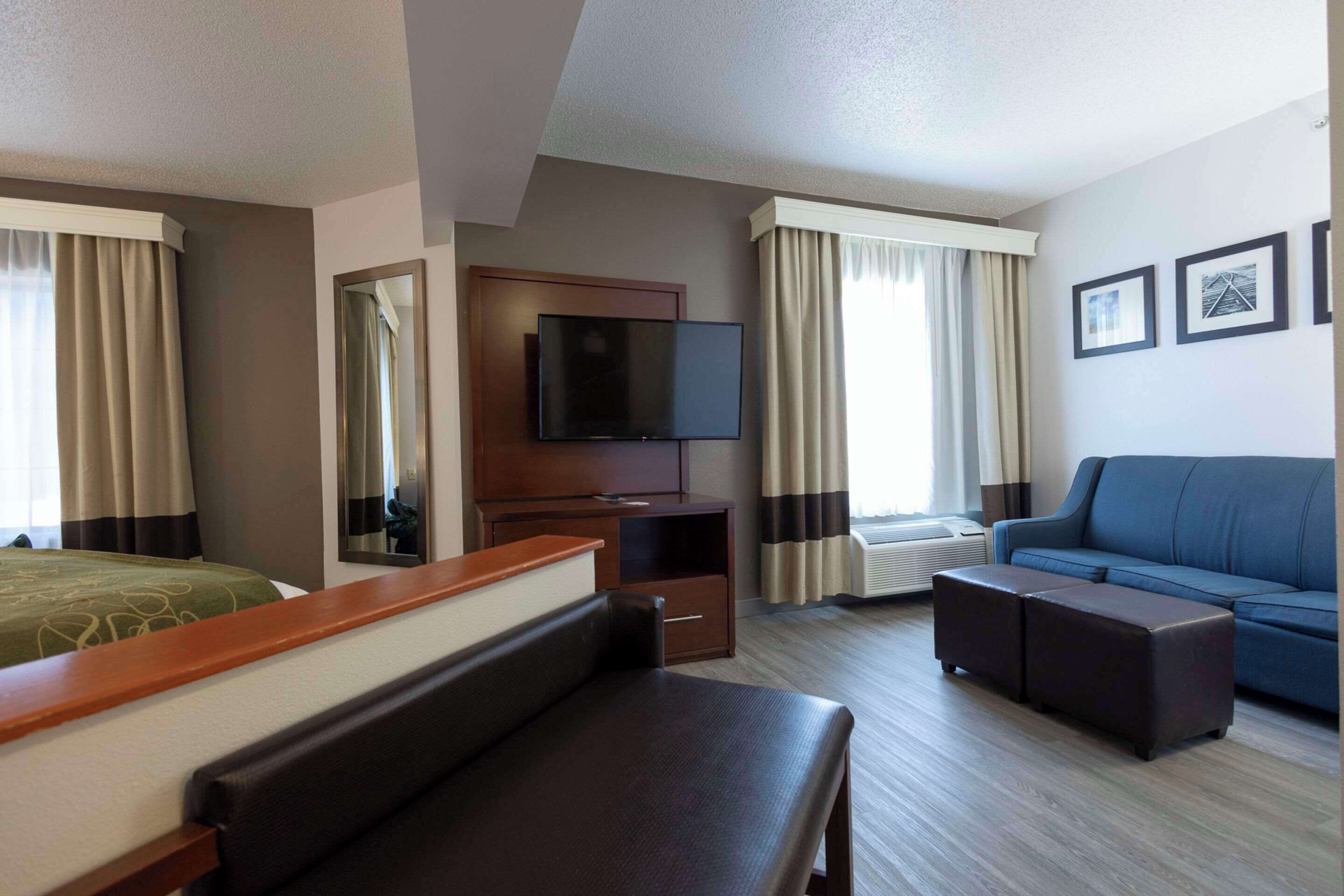 A hotel room with a bed, a desk with a flat-screen TV, a sofa, an ottoman, and a large window with curtains.