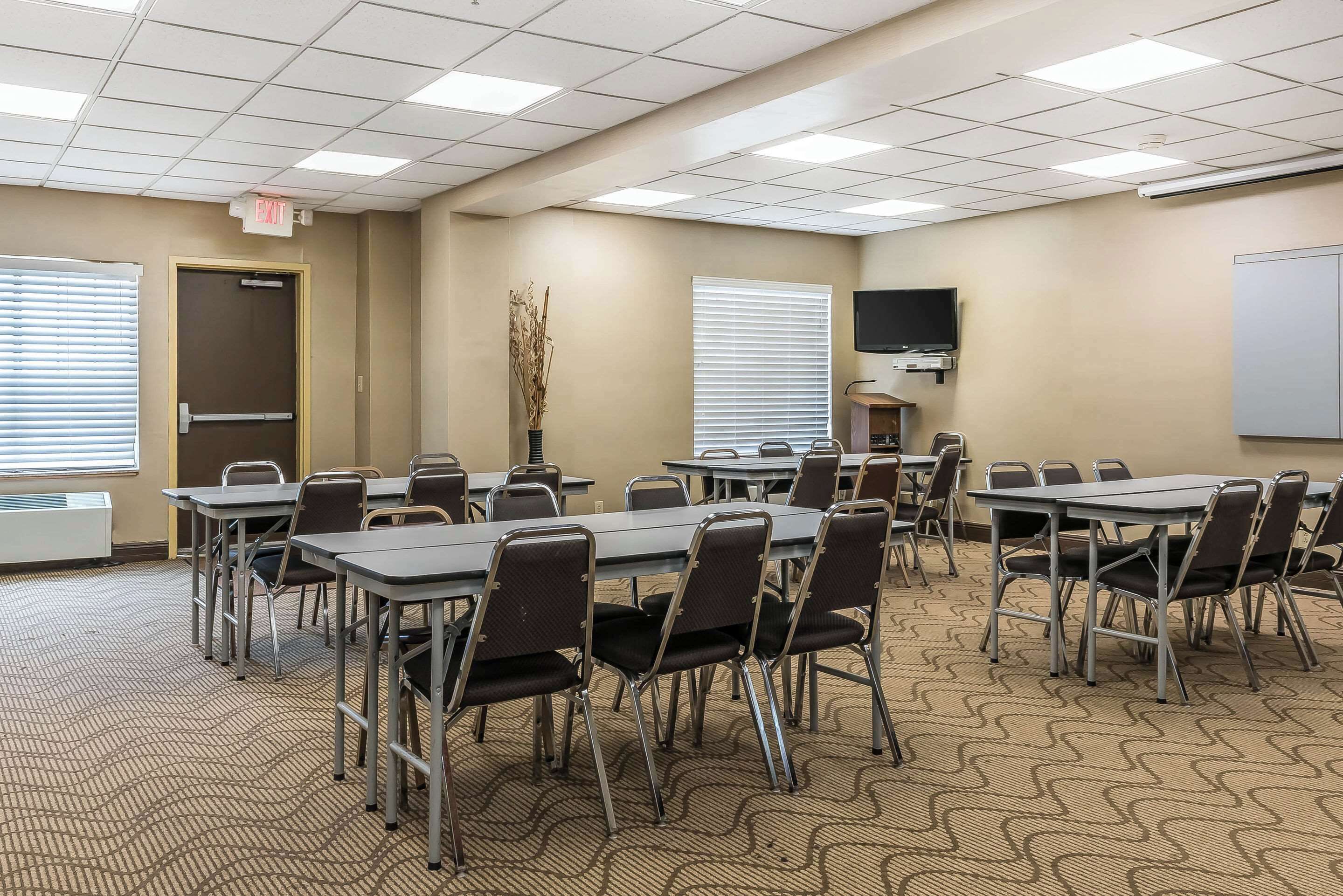 Un salón de conferencias con mesas y sillas, una puerta con letrero de salida, un televisor y una pizarra.