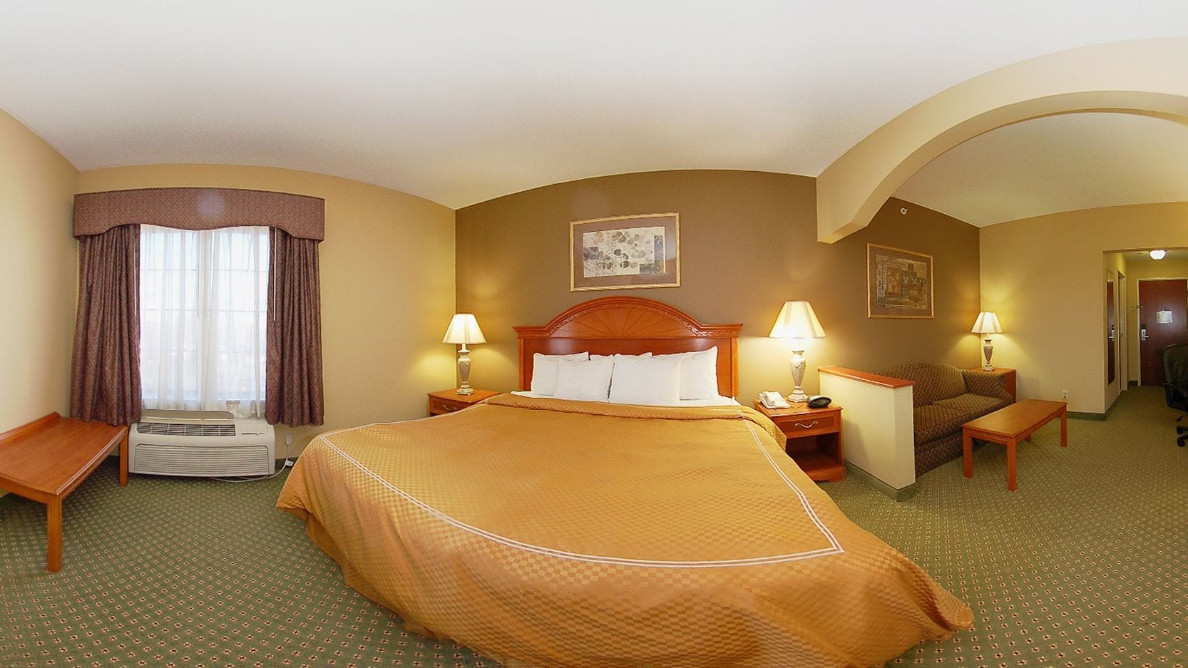 Quarto de hotel com uma cama grande, duas mesas de cabeceira com luminárias, uma cadeira, uma escrivaninha e um aparelho de ar condicionado.