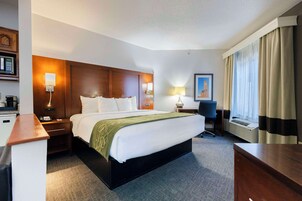 Habitación de hotel con una cama grande, mesitas de noche, un escritorio, una silla, una lámpara, un cuadro en la pared y una ventana con cortinas.