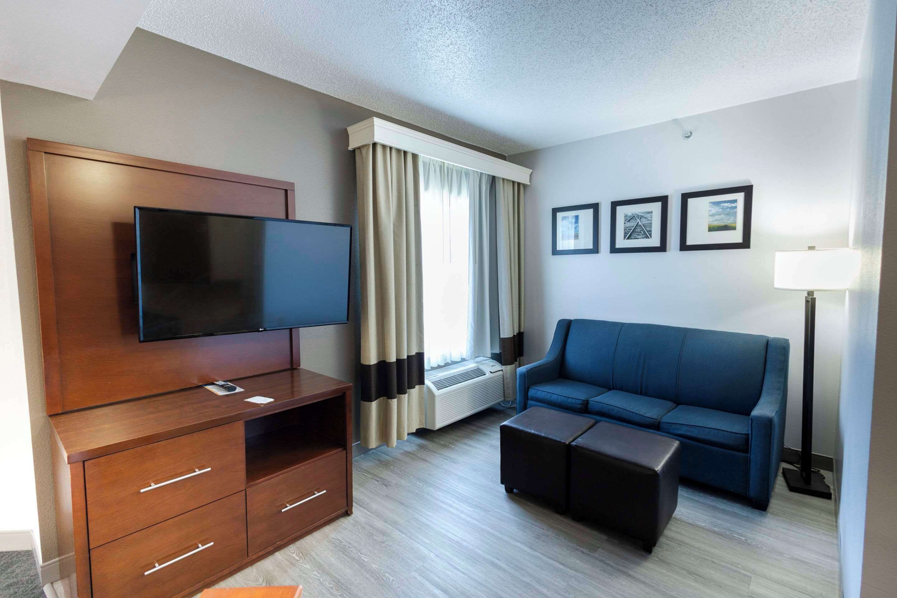 Habitación de hotel con televisor de pantalla plana, mueble de entretenimiento de madera, sofá azul, reposapiés, ventana con cortinas y tres cuadros en la pared.