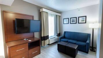 Habitación de hotel con televisor de pantalla plana, mueble de entretenimiento de madera, sofá azul, reposapiés, ventana con cortinas y tres cuadros en la pared.