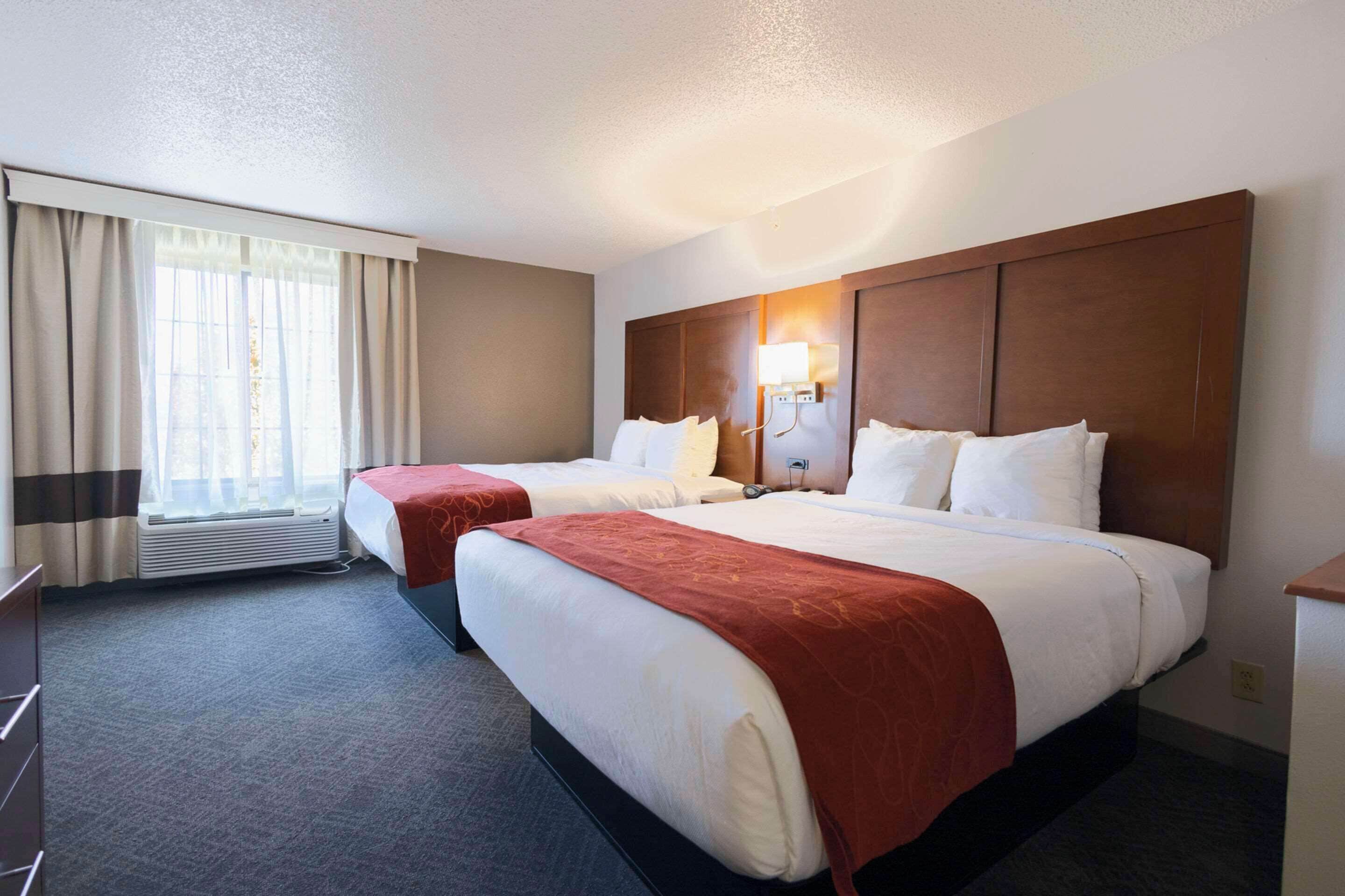 A hotel room with two beds, a large window with curtains, a wall-mounted lamp, and a desk.
