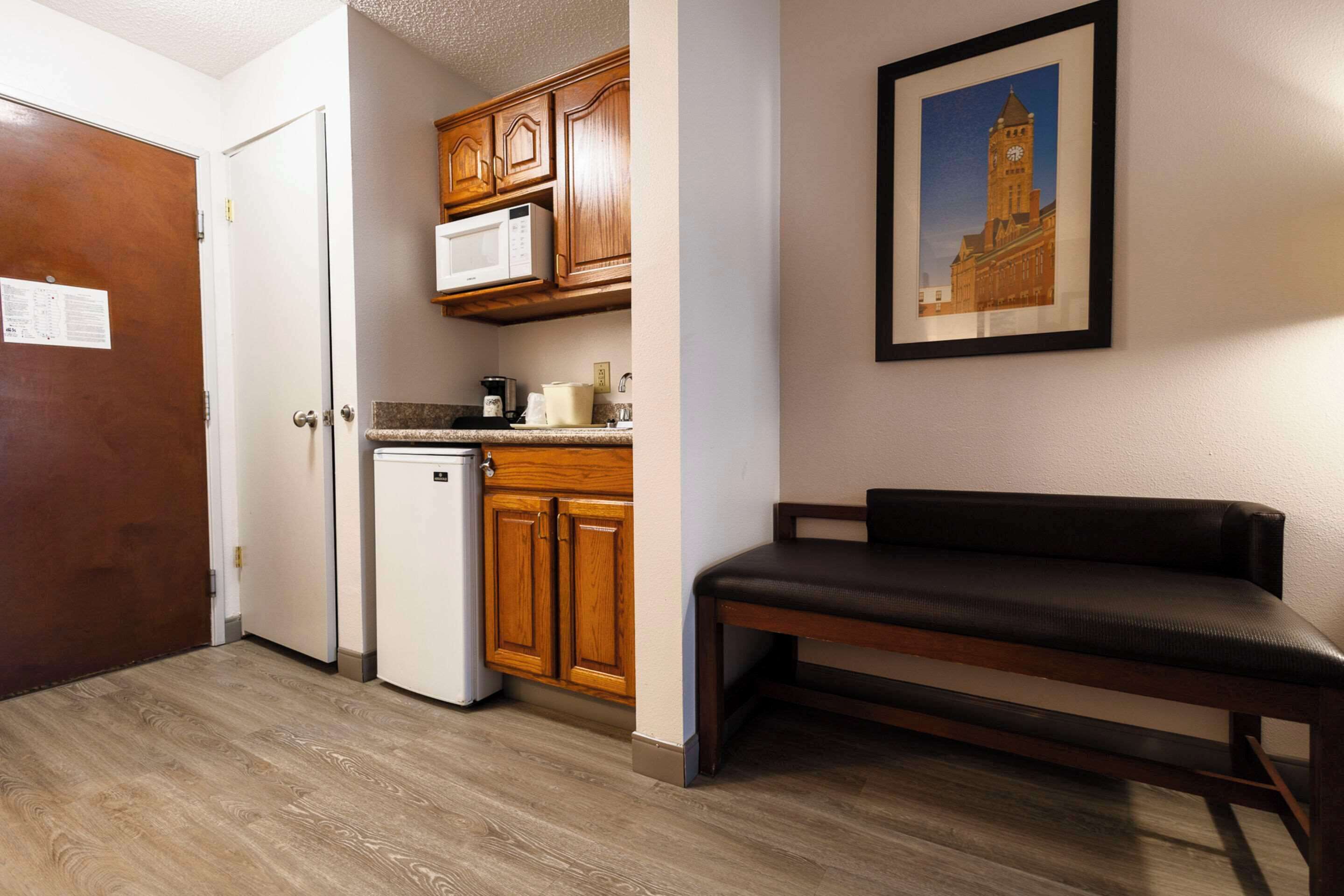 A compact kitchen with wooden cabinets, a microwave, and a refrigerator. There is a bench and a framed picture on the wall.
