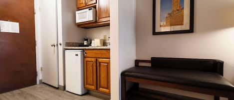 A compact kitchen with wooden cabinets, a microwave, and a refrigerator. There is a bench and a framed picture on the wall.