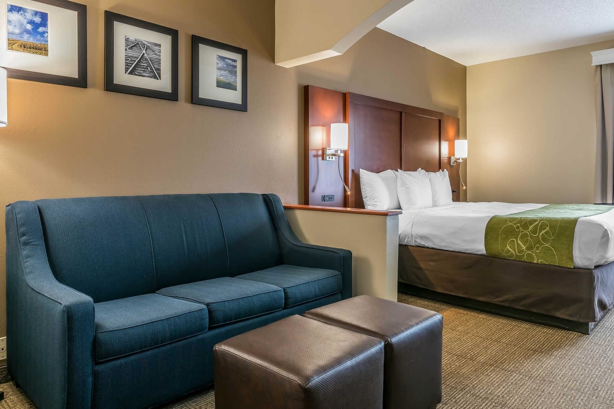 Una habitación de hotel con un sofá azul, un reposapiés marrón, una cama con una manta verde y tres cuadros en la pared.