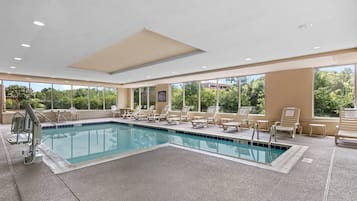 Piscina con sillas de descanso y una silla de salvavidas, rodeada de amplias ventanas con vistas a árboles.