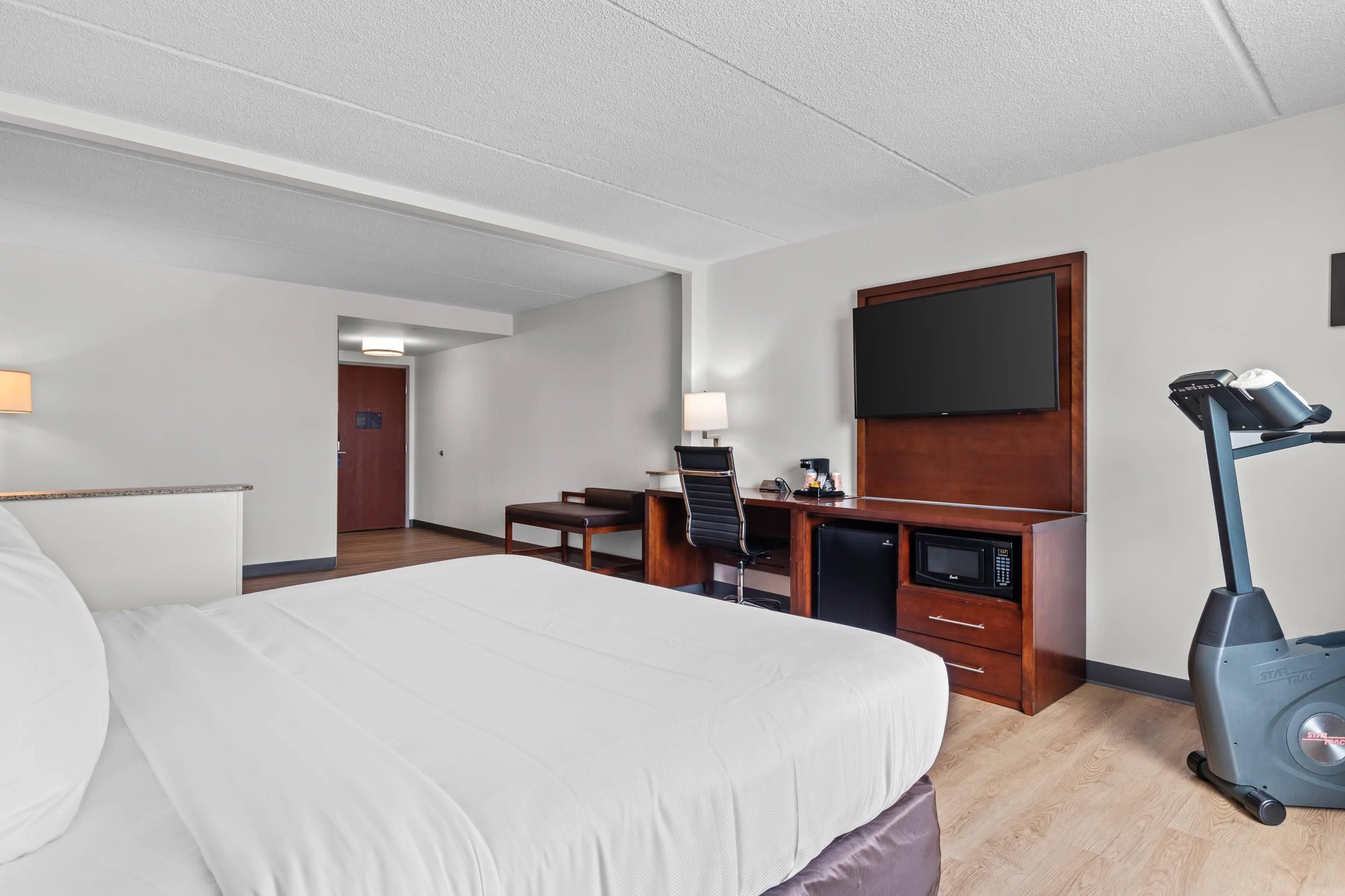 A hotel room with a bed, desk, chair, TV, microwave, and exercise bike.