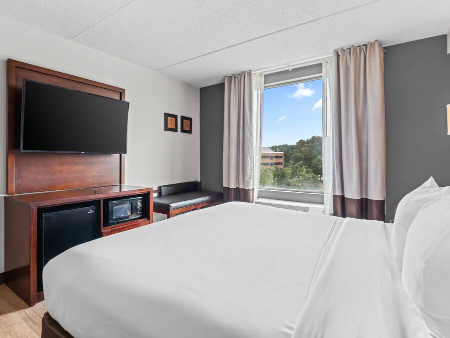 A hotel room with a bed, a flat-screen TV mounted on the wall, a small desk with a microwave, a seating area, and a window with curtains.
