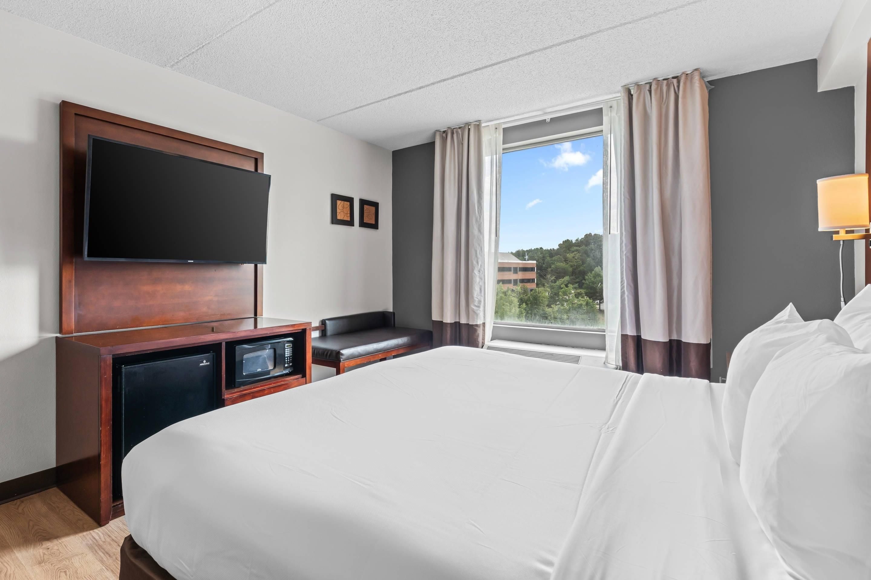 A hotel room with a bed, a flat-screen TV mounted on the wall, a small desk with a microwave, a seating area, and a window with curtains.