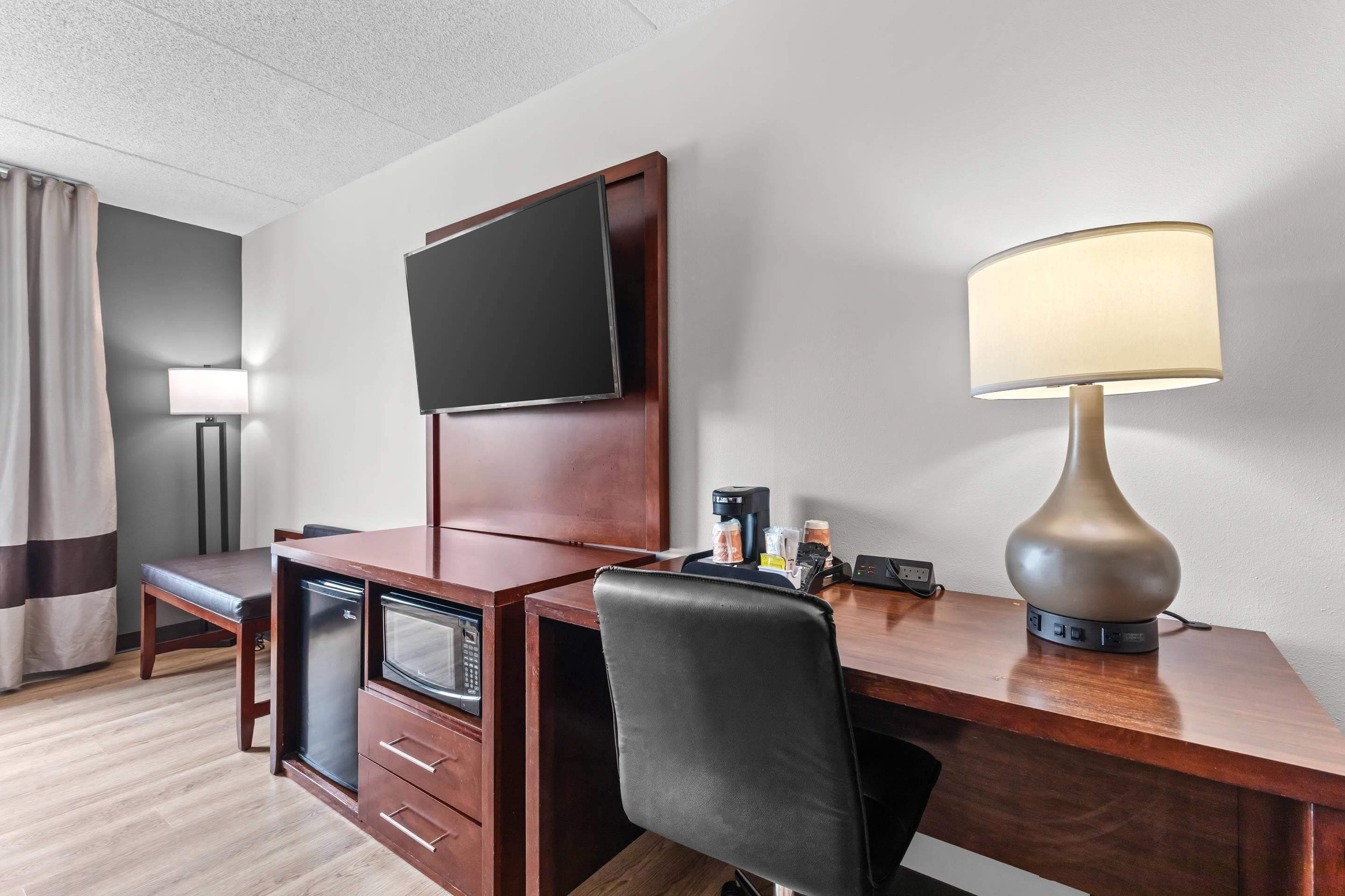 A hotel room with a wooden desk, a television mounted on the wall, a lamp, a coffee maker, and a minibar.