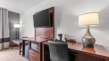 A hotel room with a wooden desk, a television mounted on the wall, a lamp, a coffee maker, and a minibar.