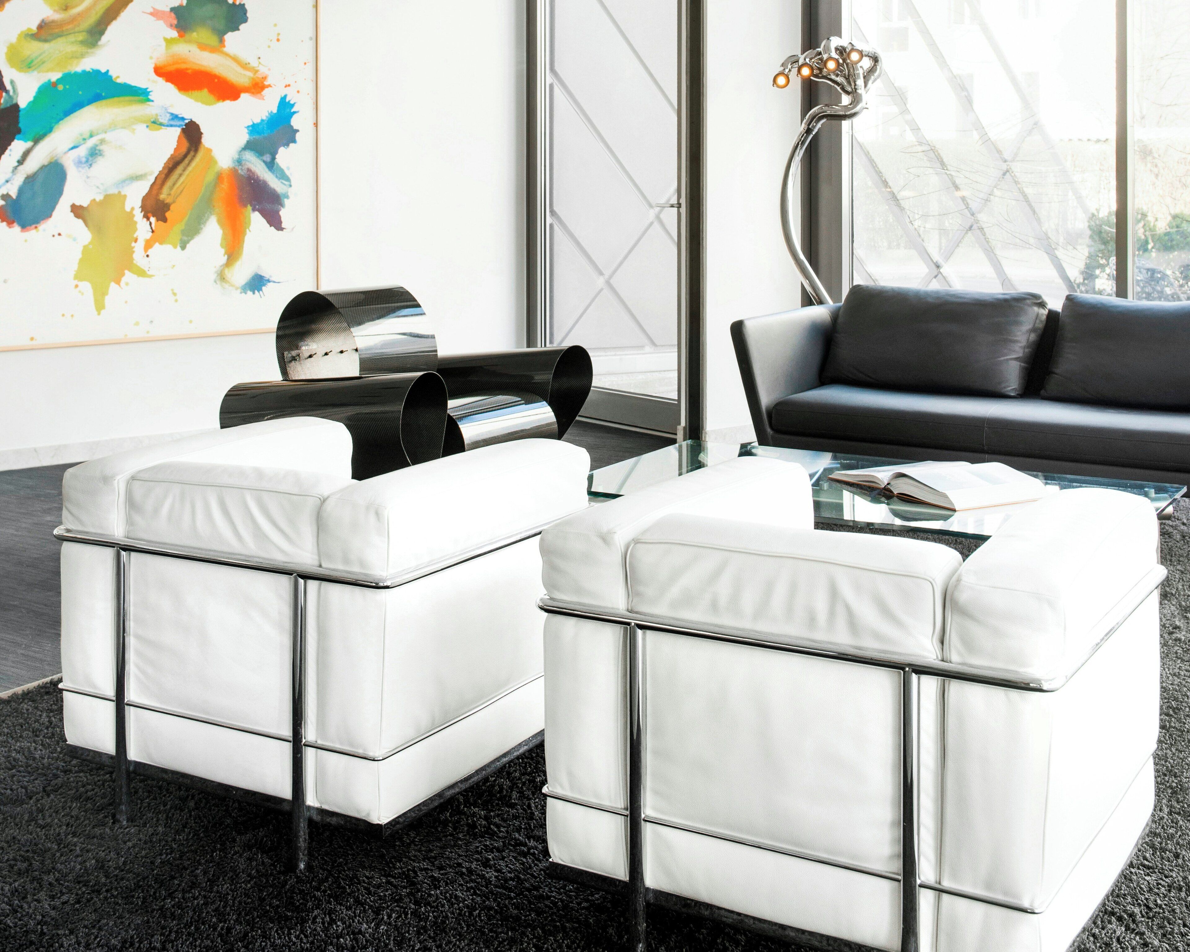 A modern living room with a black leather sofa, two white leather ottomans, a glass coffee table, and a colorful abstract painting on the wall.