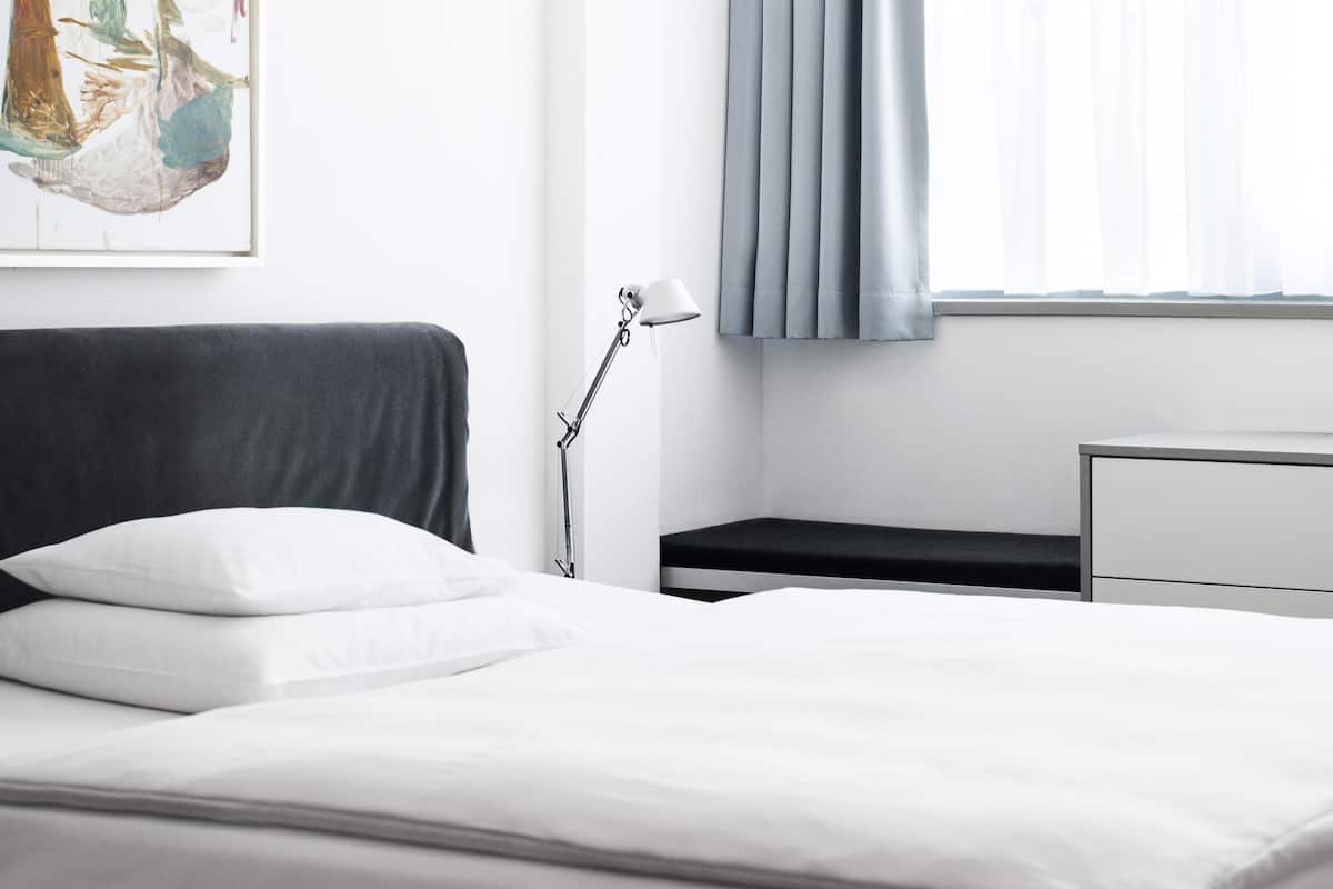 A modern hotel room with a bed, a desk, a lamp, and a window with curtains.