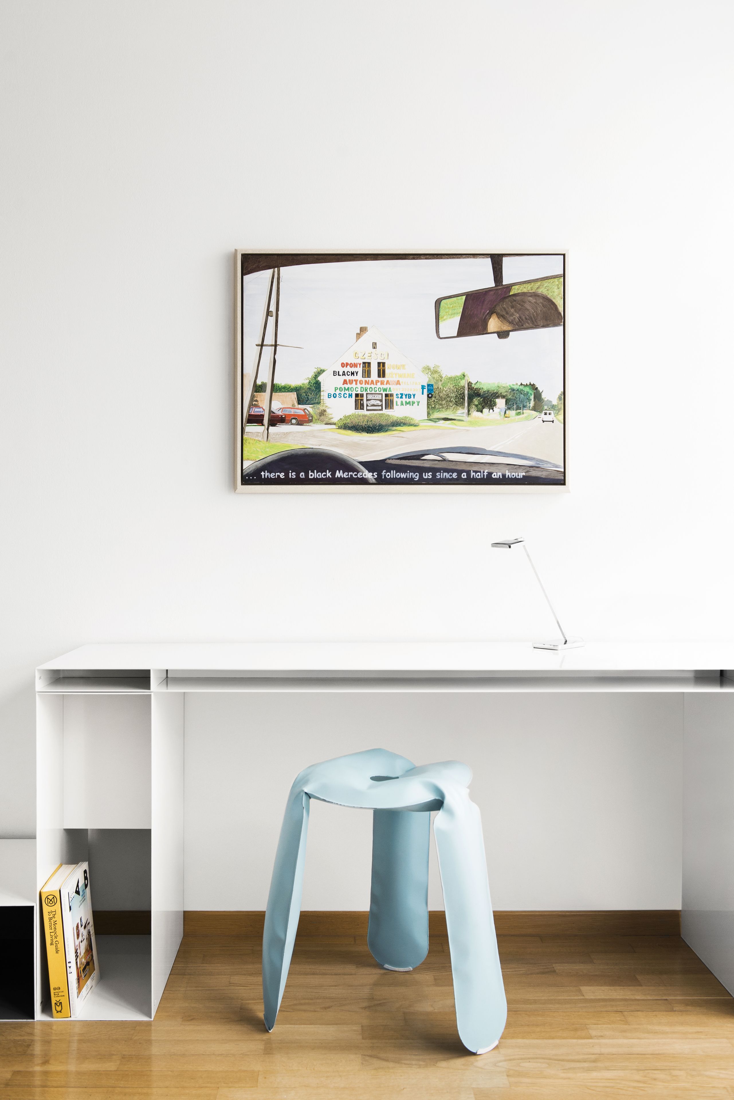 Ein moderner Arbeitsraum mit einem weißen Schreibtisch, einem blauen Hocker und einem gerahmten Bild von einer Autohaus-Werbung an der Wand.