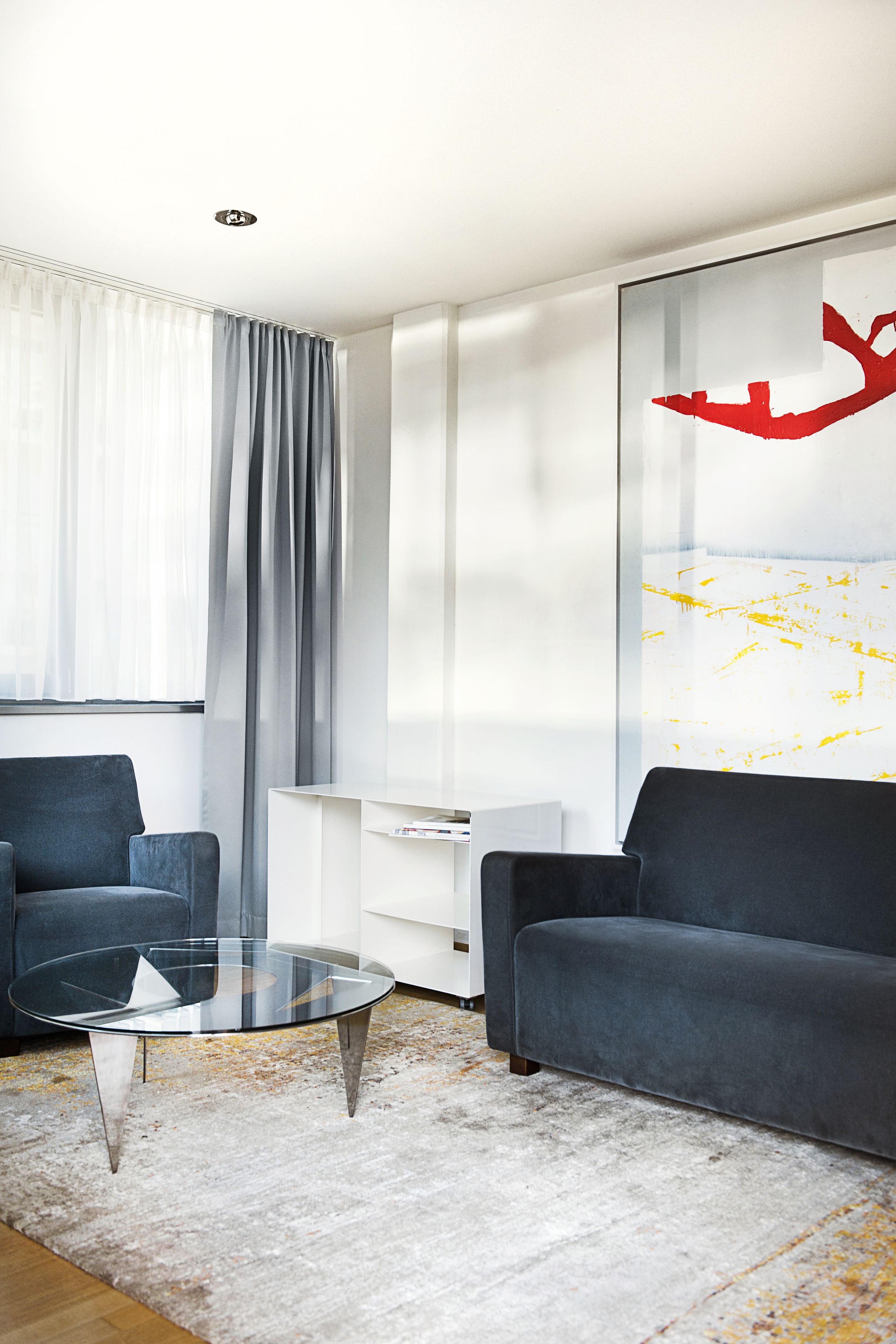 Ein modernes Wohnzimmer mit einem Glastisch, zwei Sofas, einem weißen Schrank und einem großen abstrakten Gemälde an der Wand.