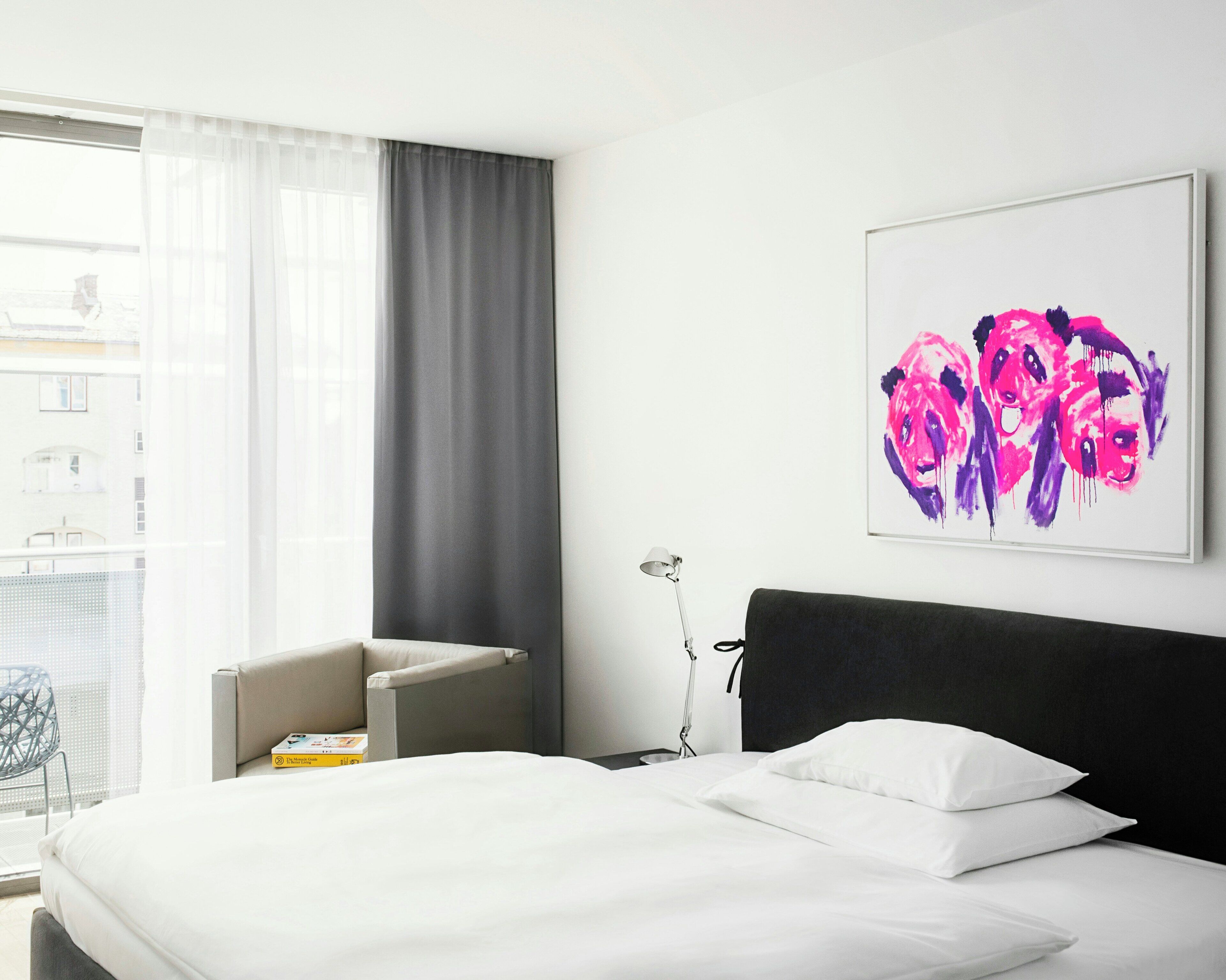 Ein Hotelzimmer mit Bett, Sessel, Nachttisch, Lampe und einem Fenster mit Vorhängen.