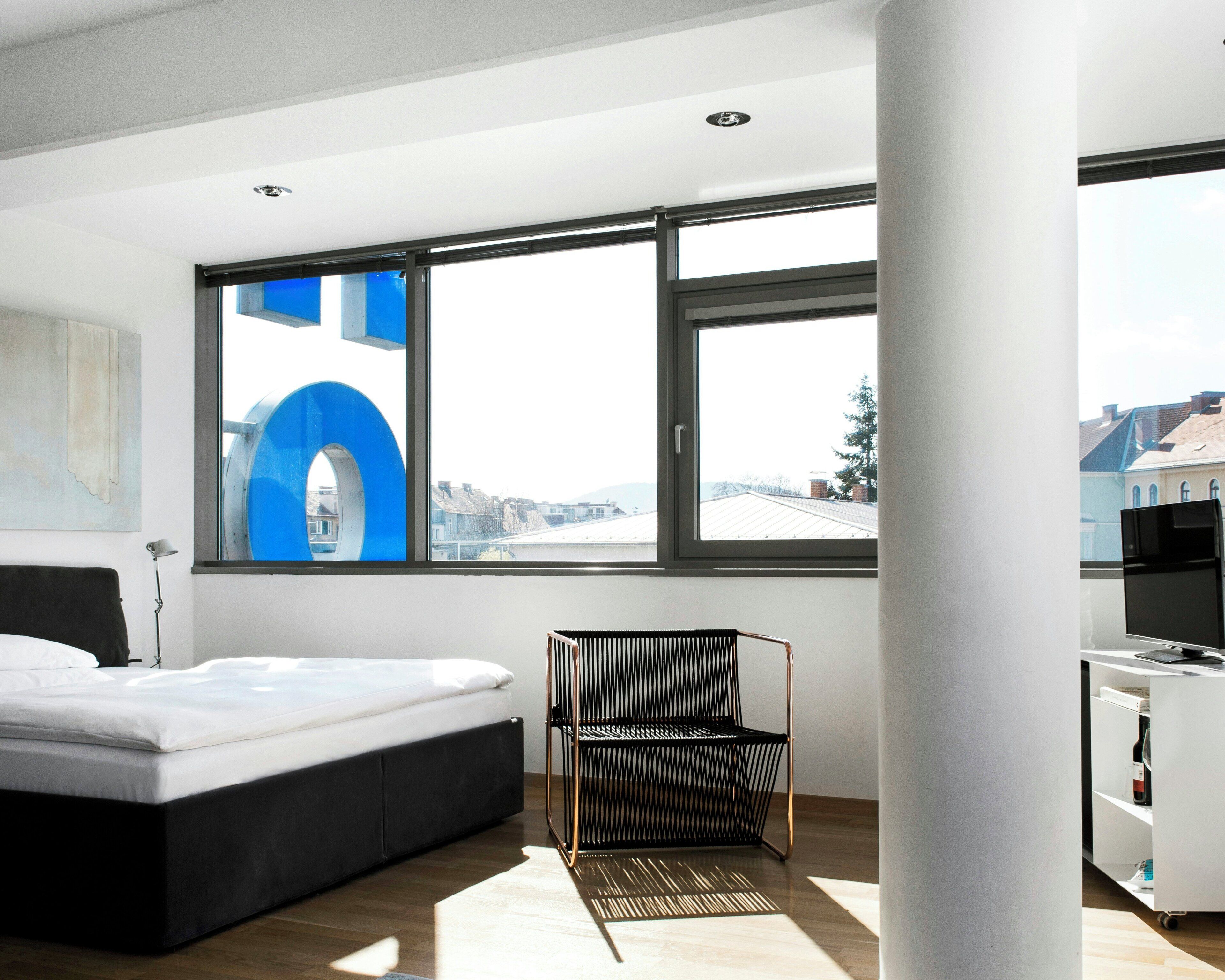 Habitación de hotel moderna con cama, una silla, un escritorio y un ventanal con vistas a la ciudad.