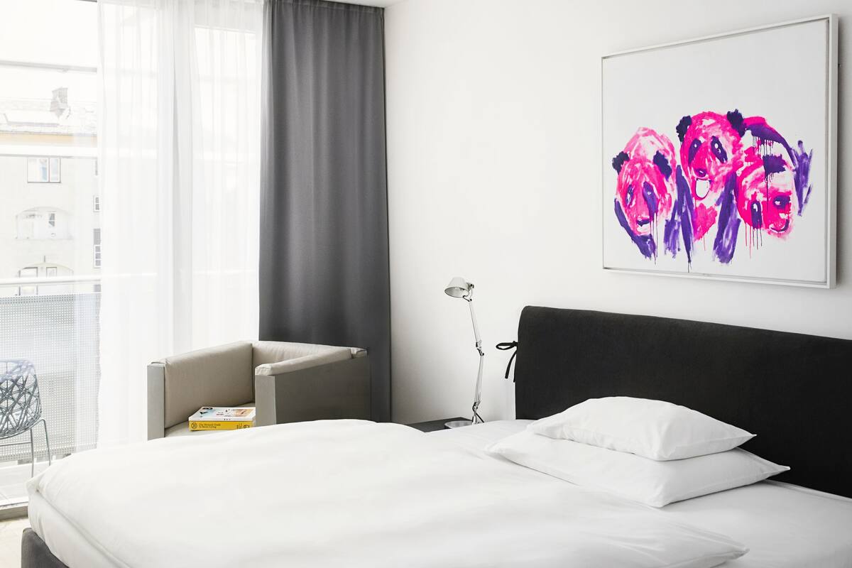 A modern hotel room with a large bed, a bedside table, a chair, and a window with curtains.