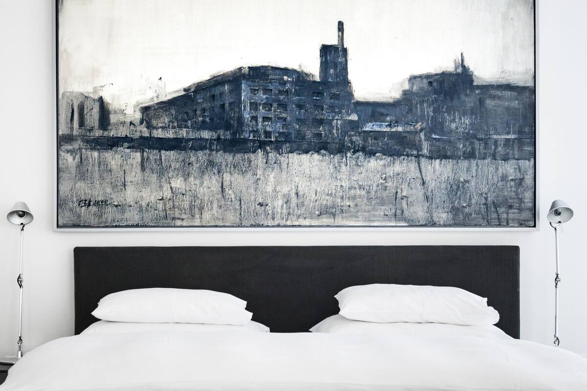 A bed with white bedding, a dark headboard, and a cityscape painting above it.