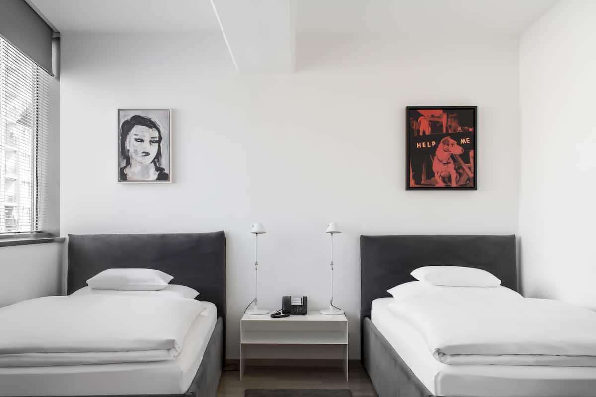 A modern hotel room with two single beds, a bedside table with a lamp, a phone, and two wall-mounted artworks.