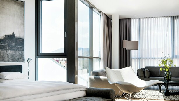Quarto de hotel moderno com uma cama grande, uma cadeira branca, um sofá, uma mesa de centro e vista para a cidade.