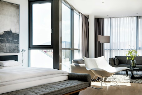 A modern hotel room with a large bed, a white chair, a sofa, a coffee table, and a view of the city.