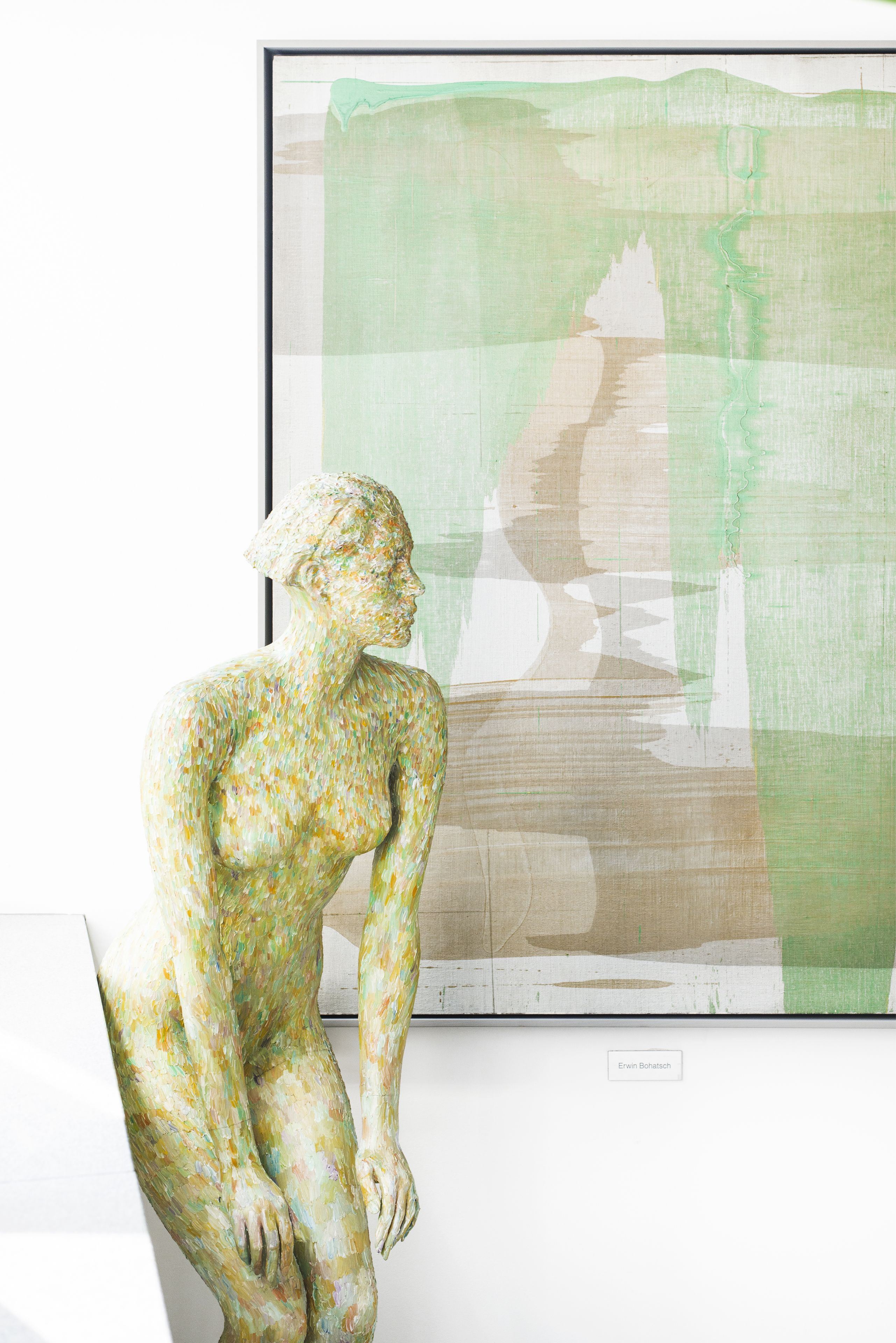 A sculpture of a seated figure in a gallery with a framed painting in the background.