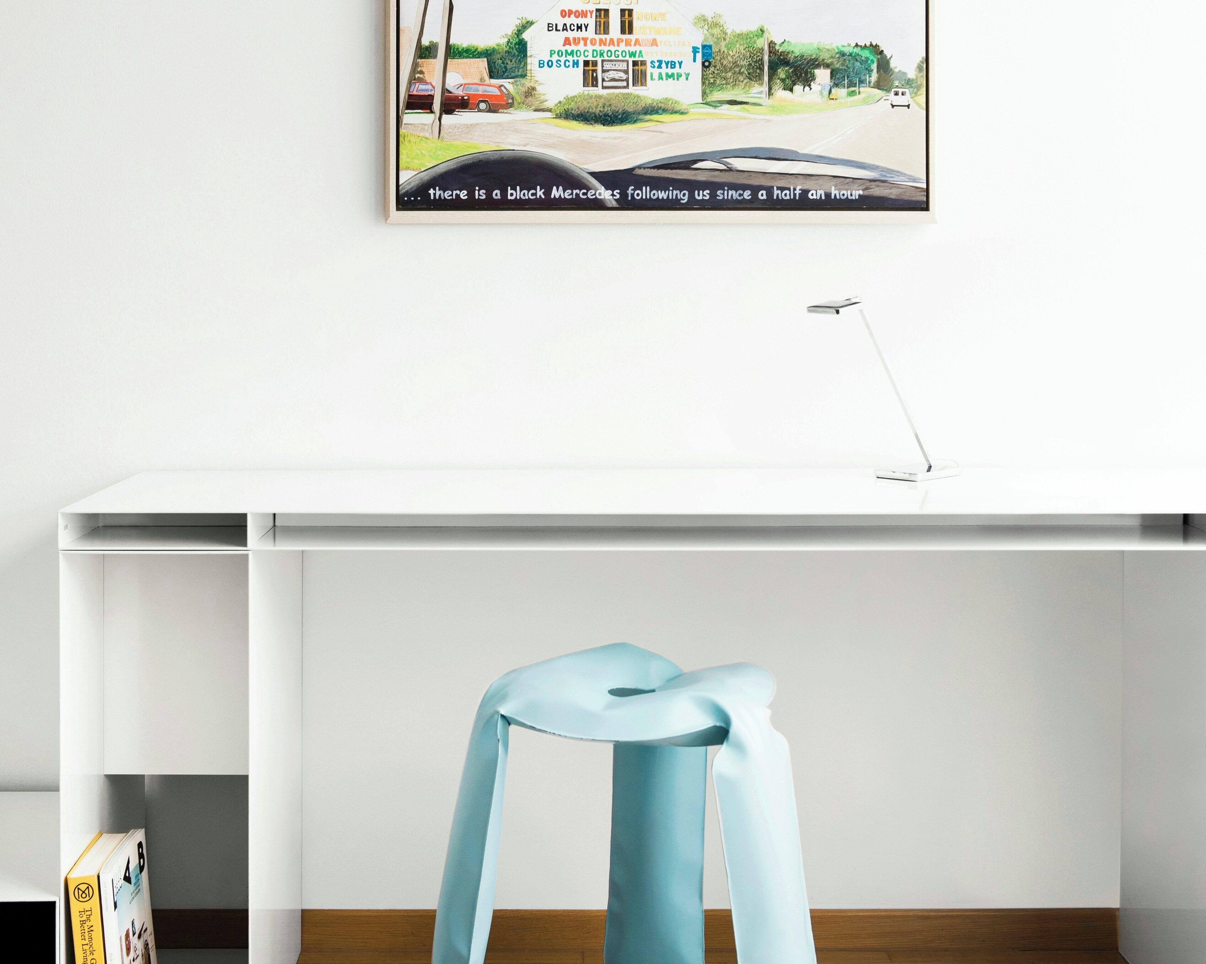 A white desk with a blue chair, a framed picture on the wall, and a lamp on the desk.