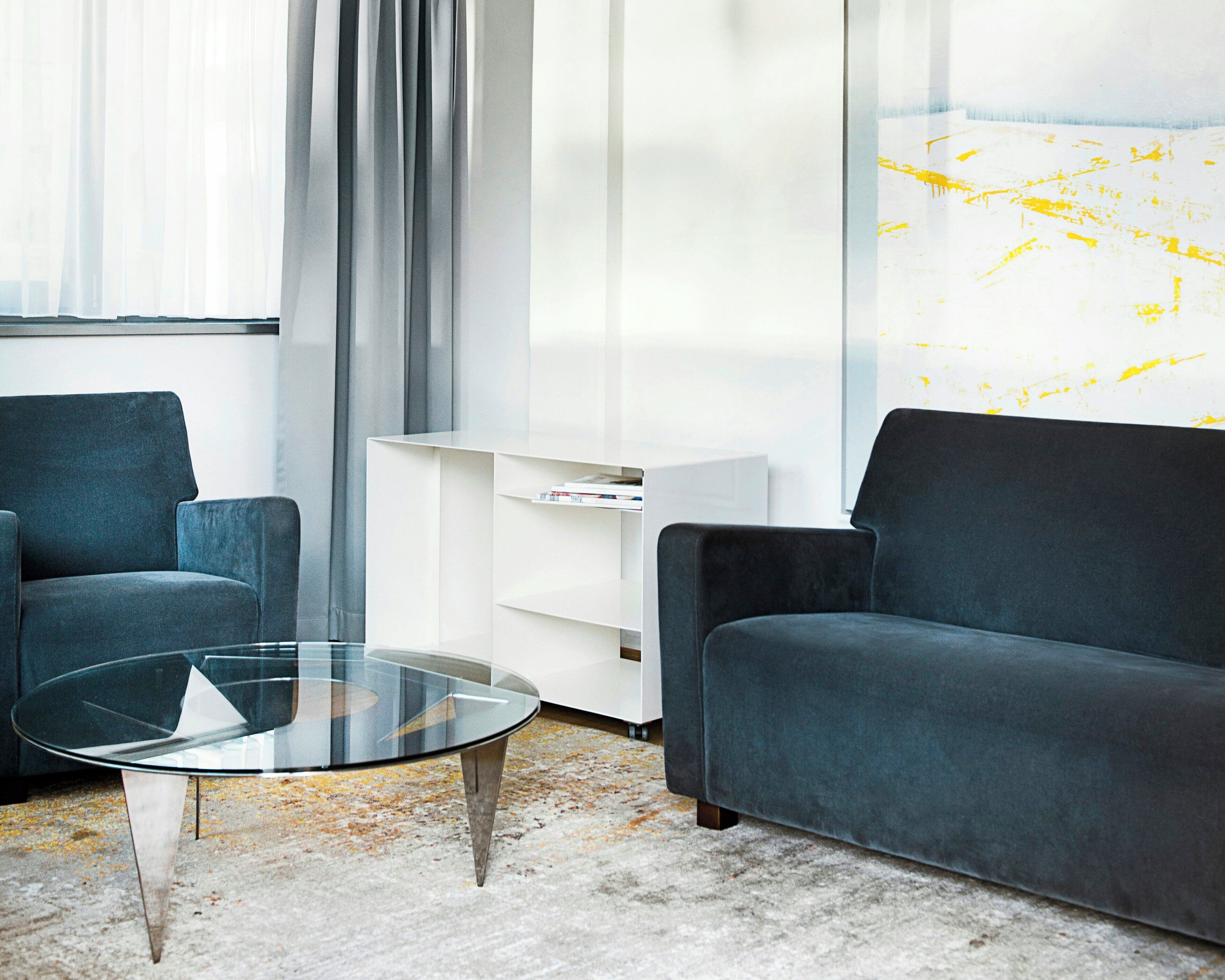A modern living room with a glass coffee table, two dark sofas, a white shelving unit, and a large window with curtains.