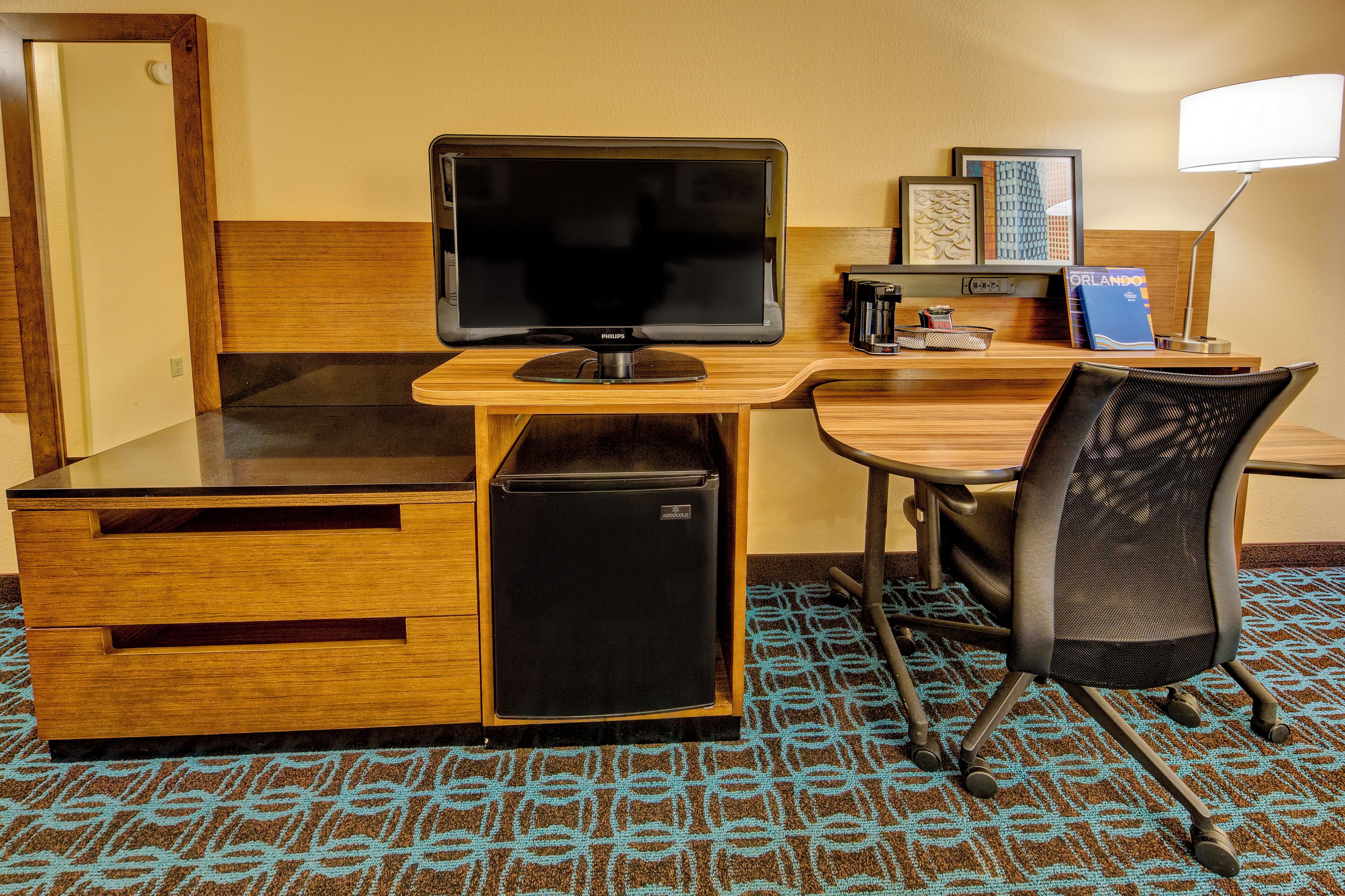 Habitación de hotel con un escritorio de madera, un monitor negro, un refrigerador pequeño, una silla de malla negra y una alfombra estampada.
