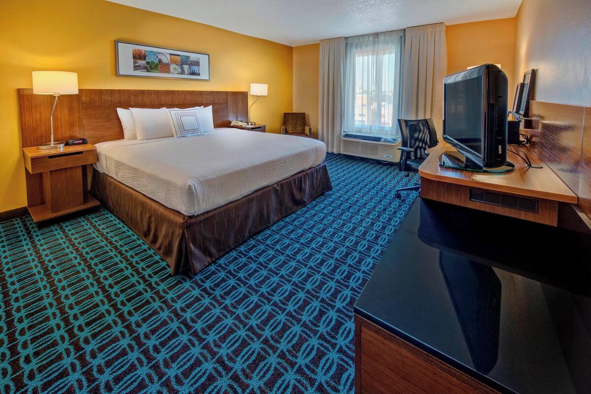 A hotel room with a large bed, a desk with a flat-screen TV, a chair, a nightstand, and a wall-mounted picture.