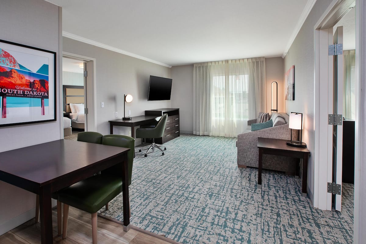 A hotel room with a dining table, a sofa, a desk with a chair, a TV mounted on the wall, and a large window with curtains.