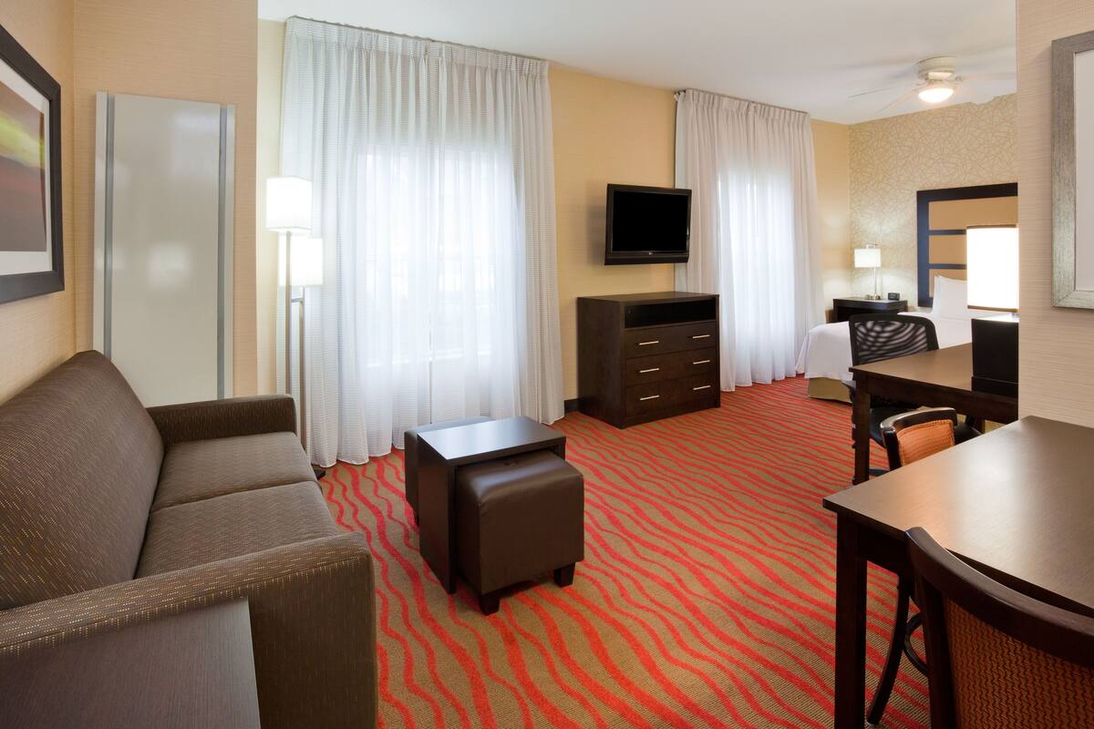 A hotel room with a sofa, a small ottoman, a TV, a desk with a chair, and a patterned carpet.