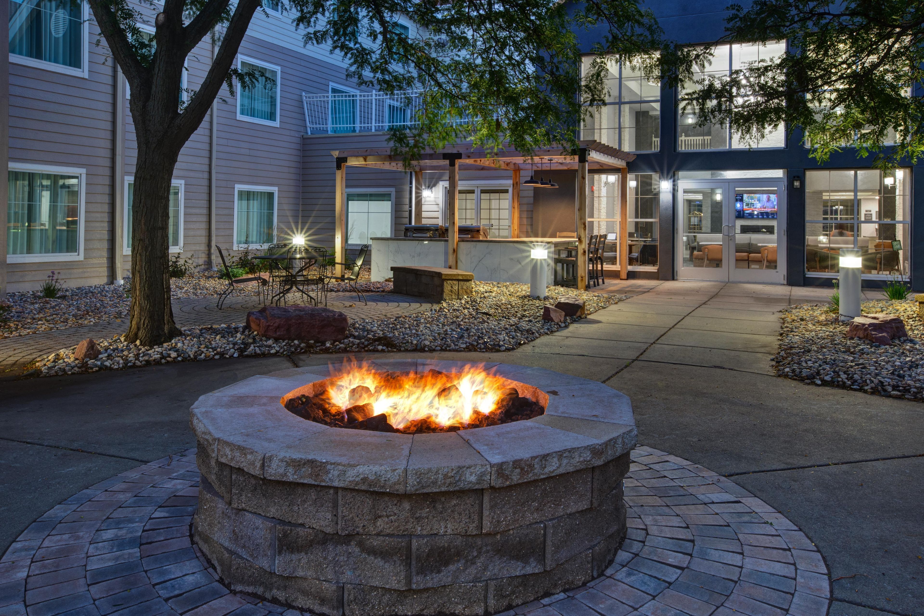 An outdoor seating area with a fire pit, surrounded by a stone wall and a pathway leading to a building entrance.