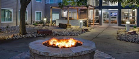 An outdoor seating area with a fire pit, surrounded by a stone wall and a pathway leading to a building entrance.
