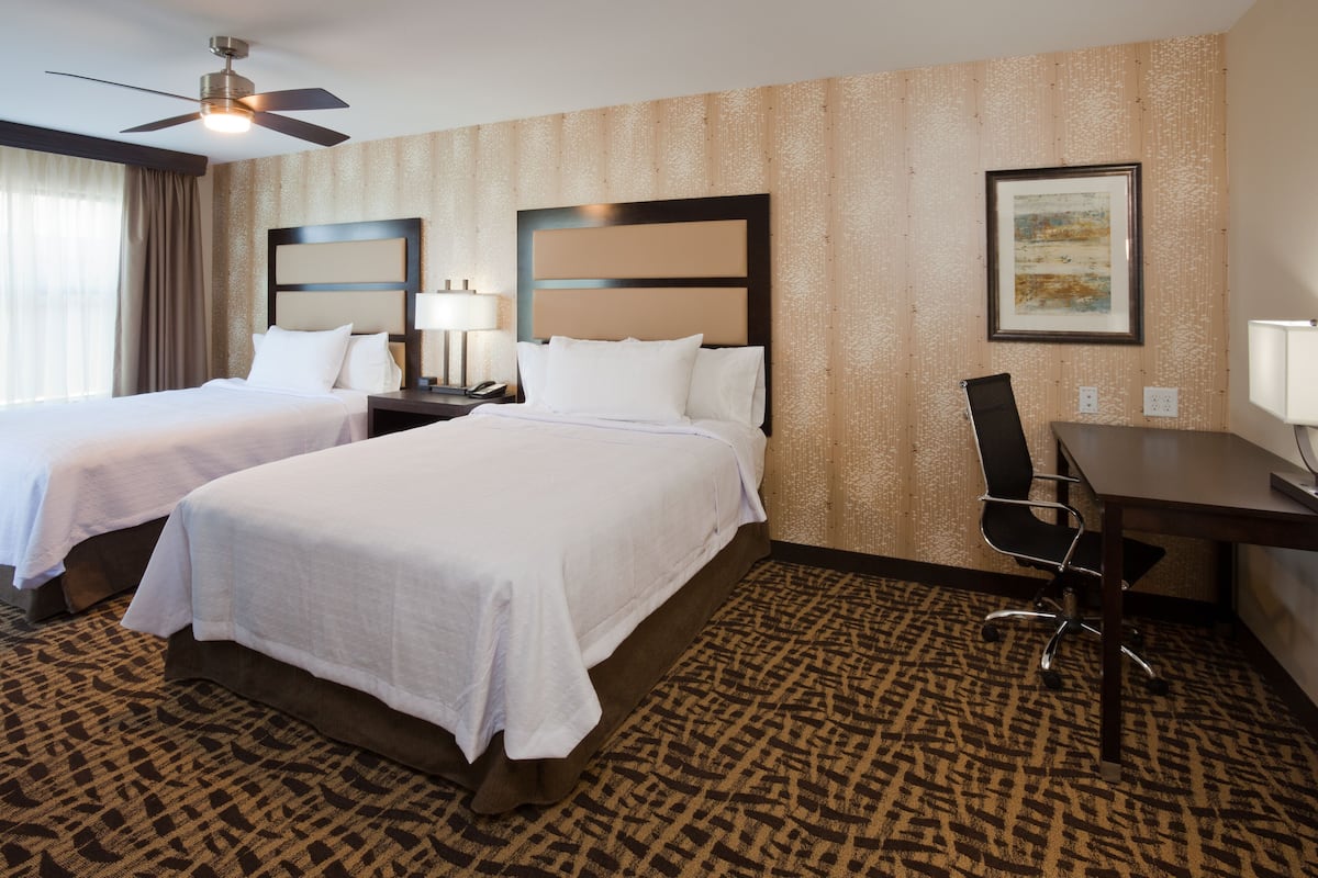 A hotel room with two beds, a desk, a chair, a ceiling fan, and a framed picture on the wall.