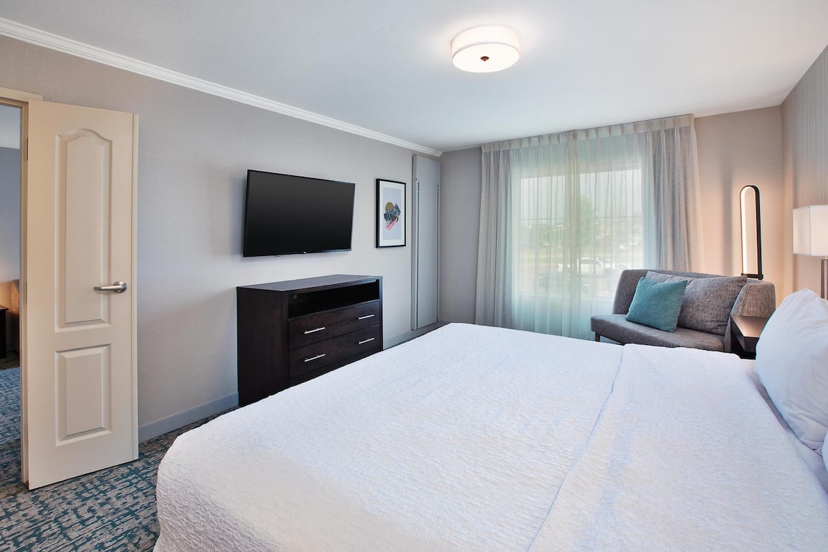 A hotel room with a large bed, a flat-screen TV mounted on the wall, a small seating area with a couch and pillows, and a door leading to another room.