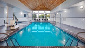 An indoor swimming pool with a wooden ceiling, clear glass walls, and a seating area with tables and chairs.