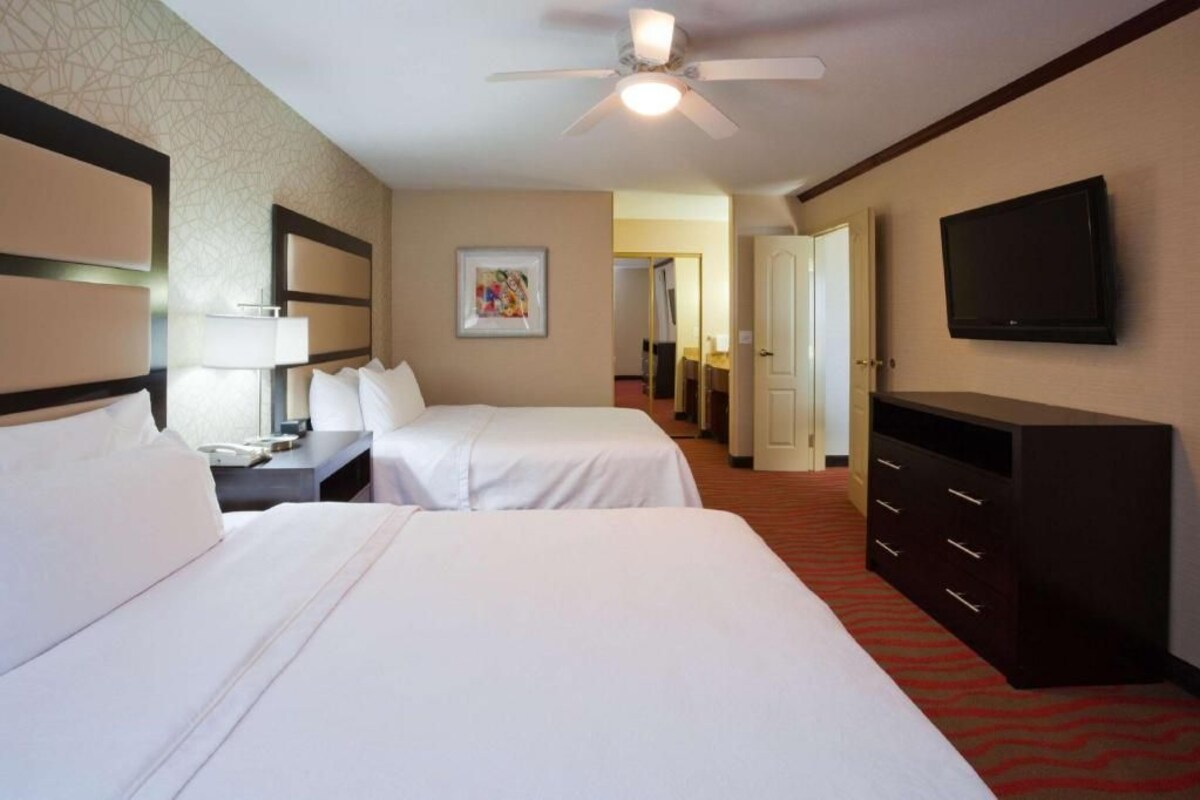 Une chambre d’hôtel avec deux lits, un bureau, une commode, une télévision et un ventilateur de plafond.